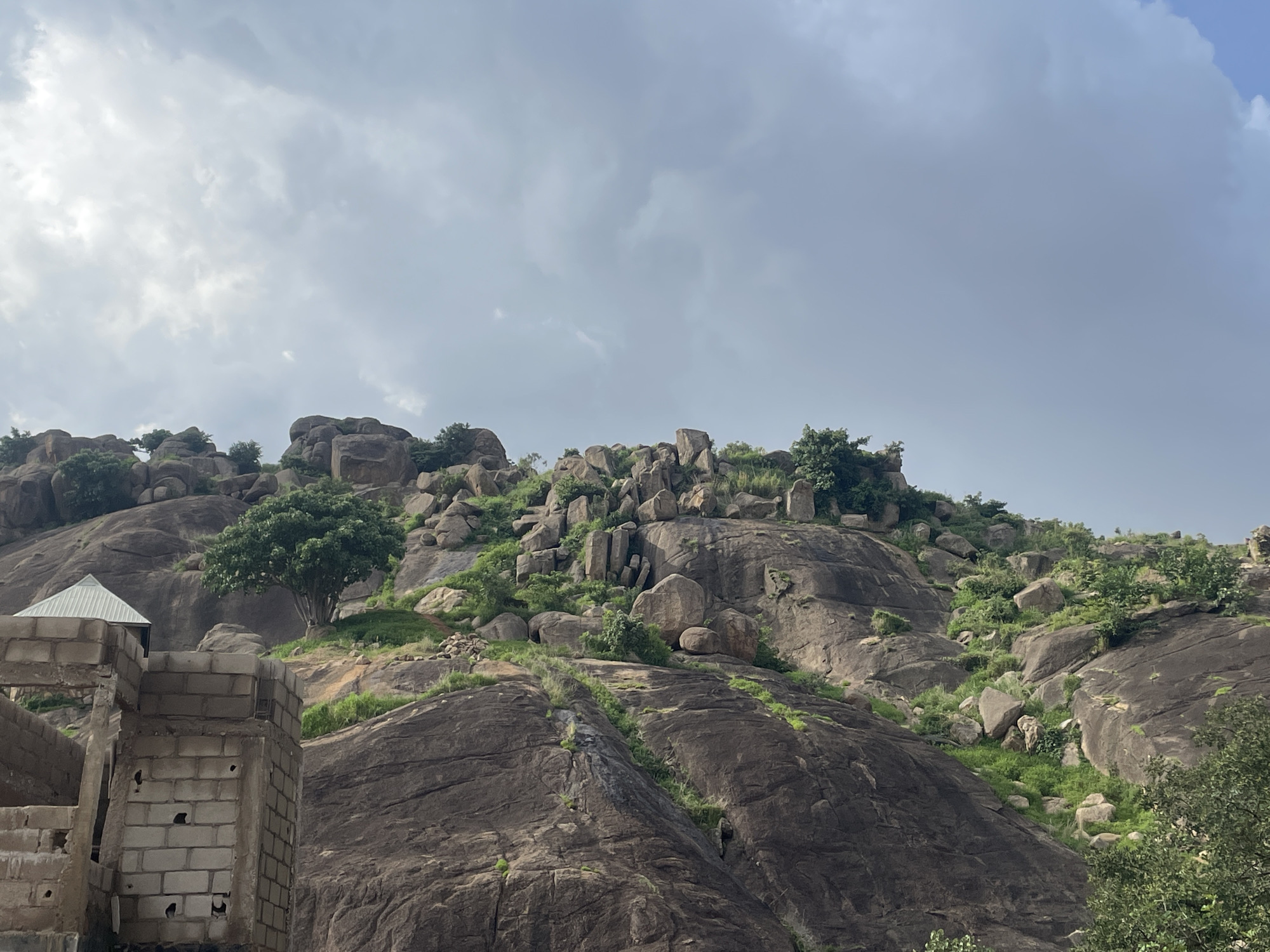 The image depicts a rugged, rocky hillside covered in sparse green vegetation and a few scattered trees. The terrain is characterized by large boulders and weathered rock formations. In the foreground, to the left, a partially constructed building made of concrete blocks is visible. The building has an unfinished appearance, with exposed brickwork and a metal roof. The sky above is overcast with heavy, gray clouds. The overall scene suggests a natural, somewhat undeveloped landscape with evidence of human construction.