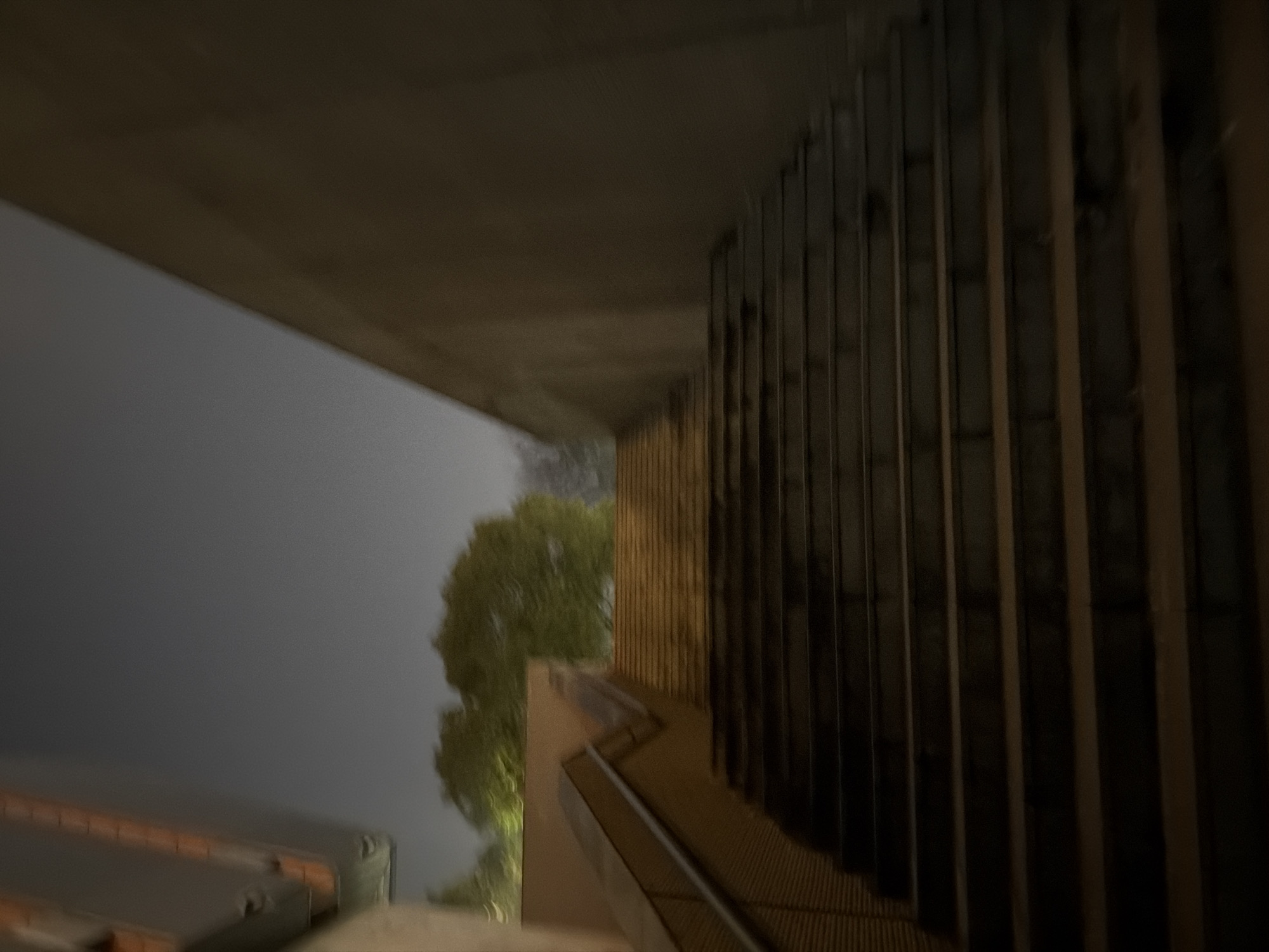 A blurry image shows an outdoor staircase in Jiangning District, China. The scene is dominated by a large concrete structure overhead, possibly a bridge or overpass. The staircase is made of a textured material, possibly metal grating, and features a handrail. To the right of the staircase is a wall with vertical slats or panels. In the background, through an opening, are trees and a gray sky. The image suffers from motion blur and low light conditions.