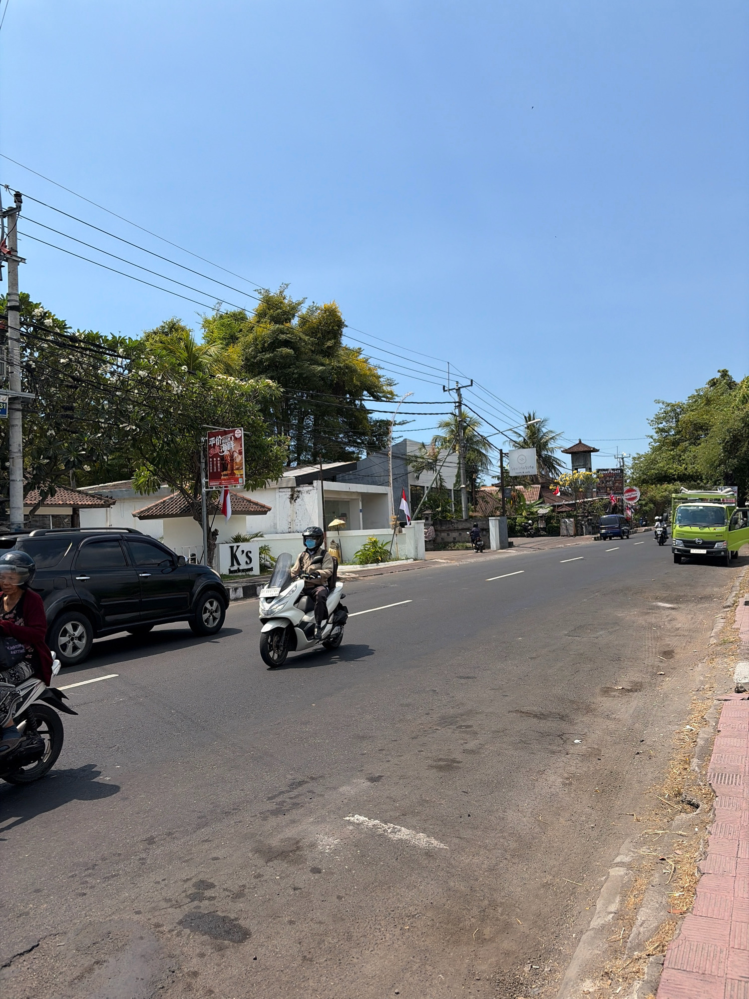 The image depicts a sunny daytime scene on a paved road in Banjar, Indonesia. A black SUV is parked on the left side of the road, with a motorcycle and rider visible in the foreground to the left, and another motorcycle with a rider in the middle of the frame moving away from the viewer. A person is riding a white scooter in the middle of the road, wearing a helmet and a light-colored jacket. On the right side of the road, a bright green truck is parked. The road is bordered by a sidewalk with red tiles on the right. Buildings with traditional Indonesian architecture and lush green trees line the sides of the road, with power lines stretching across the sky. A red and white Indonesian flag is visible on a pole in front of one of the buildings. The sky is clear and blue.