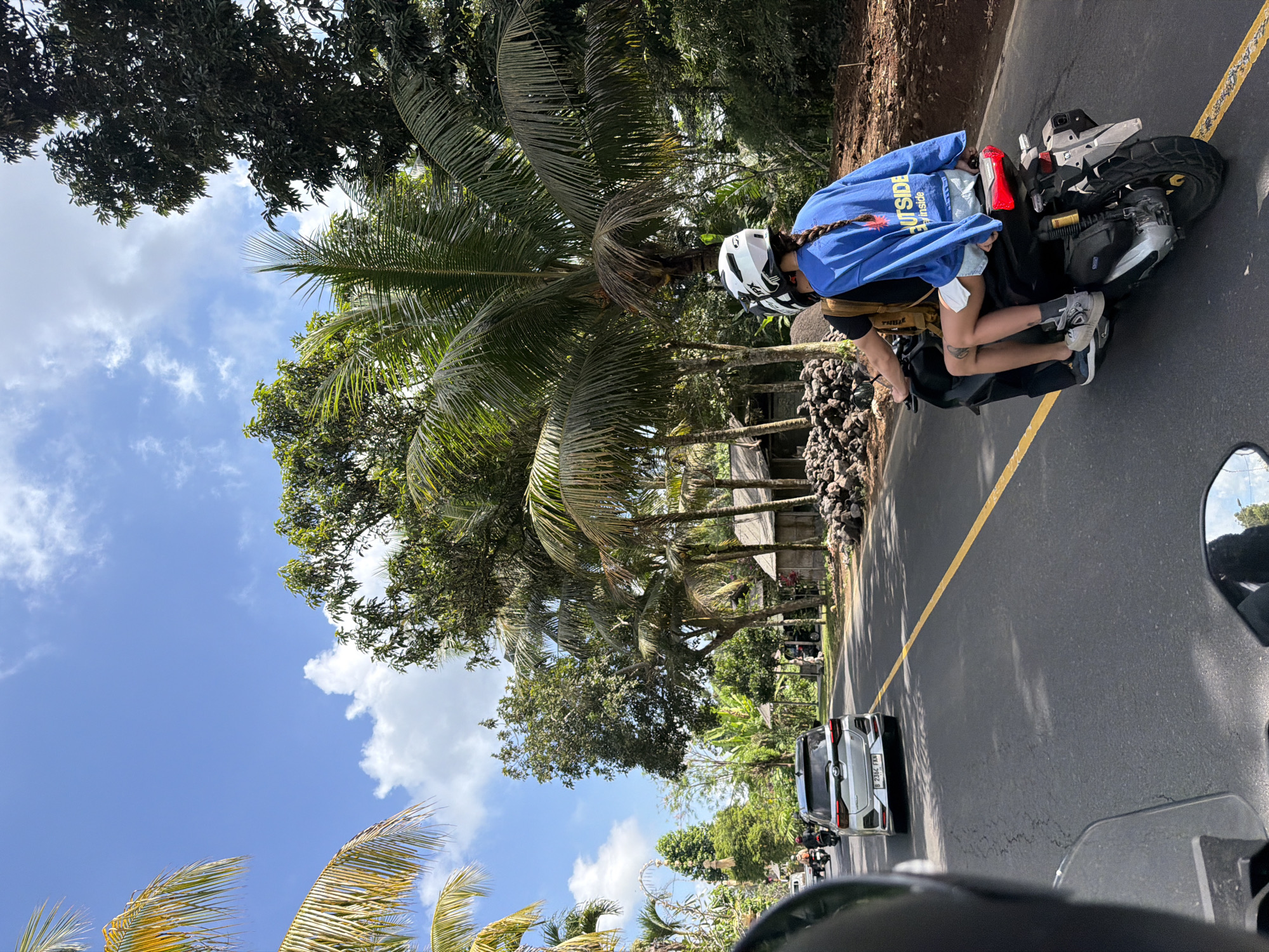 In Tampaksiring, Indonesia, on a sunny day, two people ride a scooter down a paved road. The person in front wears a white helmet and a black backpack. The person behind wears a white helmet with a braid of brown hair extending from it and a blue long-sleeved shirt that reads "OUTSIDE / inside." They are wearing denim shorts and sneakers. A silver SUV is traveling in the opposite direction. The road is lined with palm trees, lush green vegetation, and several small huts with piles of rocks. The sky is blue with scattered white clouds. The image is taken from the perspective of someone on another scooter, looking forward.