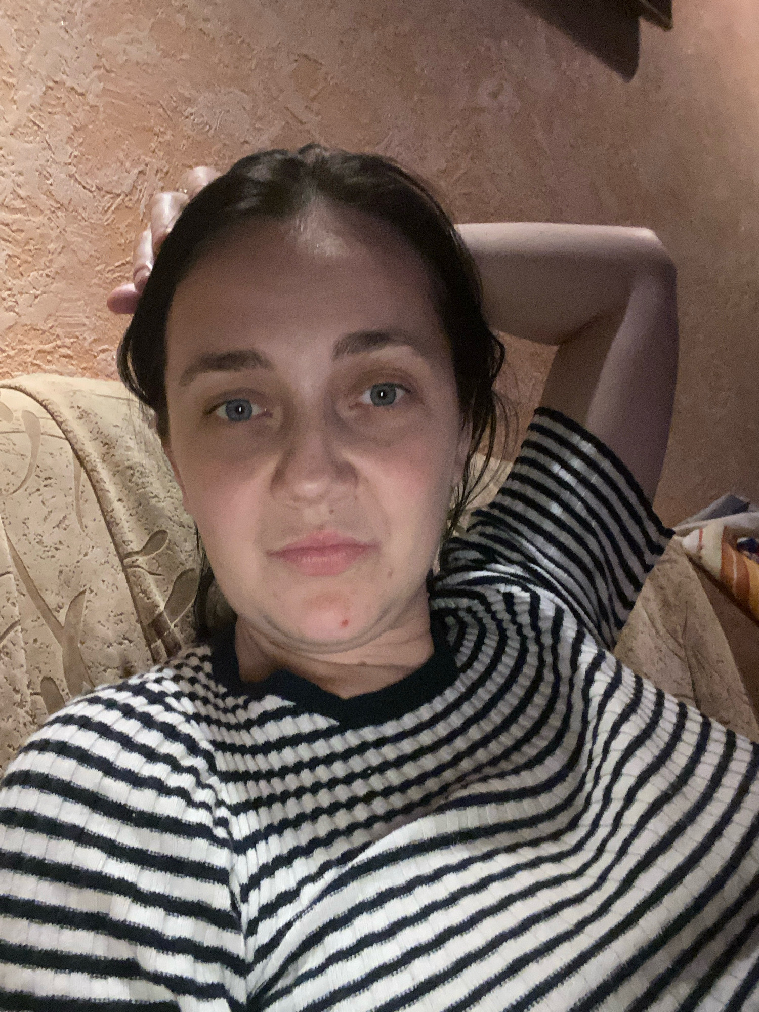 A woman with dark hair and blue eyes is lying on a couch. She is wearing a black and white striped shirt. Her left arm is raised and resting on her head. The wall behind her is a textured, light brown color. The couch is covered in a beige fabric with a floral pattern.