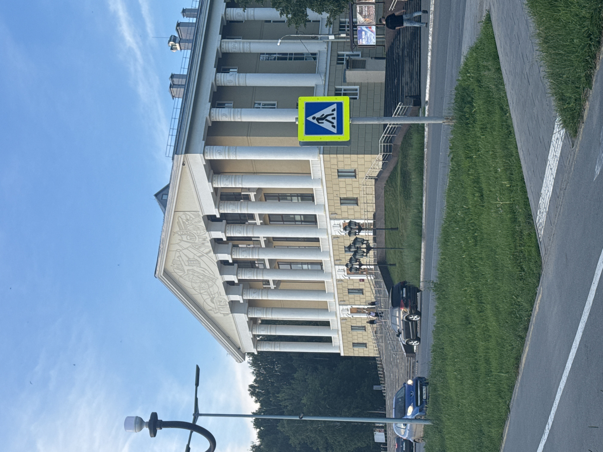 The image shows a wide, paved road in front of a large, neoclassical building in Votkinsk, Russia. The building has a prominent facade with numerous white columns and an ornate frieze. Several cars are visible on the road, including a blue hatchback and a black SUV. A pedestrian crossing sign is positioned on the right side of the road. Green grass and trees line the area between the road and the building. The sky is a clear blue with a few wisps of clouds. The overall scene depicts a public space in a city, likely during daylight hours.
