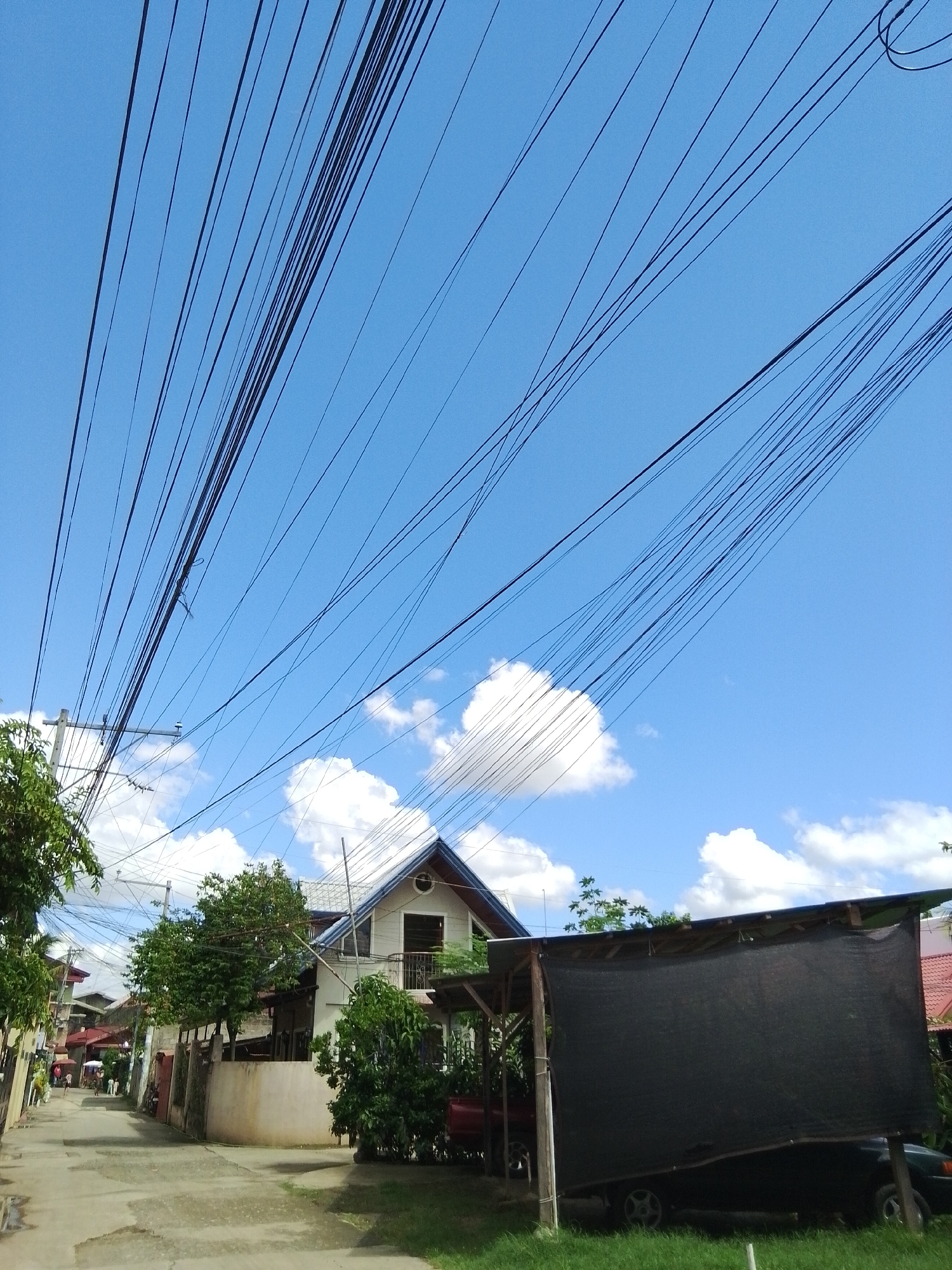 The image depicts a residential street in Davao City, Philippines, characterized by an overcast sky and a network of overhead power lines. In the foreground, a portion of a property with a black tarp-covered structure housing a dark-colored vehicle is visible on the right. Adjacent to this, a concrete wall and lush greenery border a paved street. The street extends into the distance, lined with houses and trees, with a few pedestrians visible. A prominent white house with a pitched roof stands out in the mid-ground. The overall scene suggests a typical urban or suburban environment within Davao City, with the prominent power lines and dense vegetation being characteristic features.