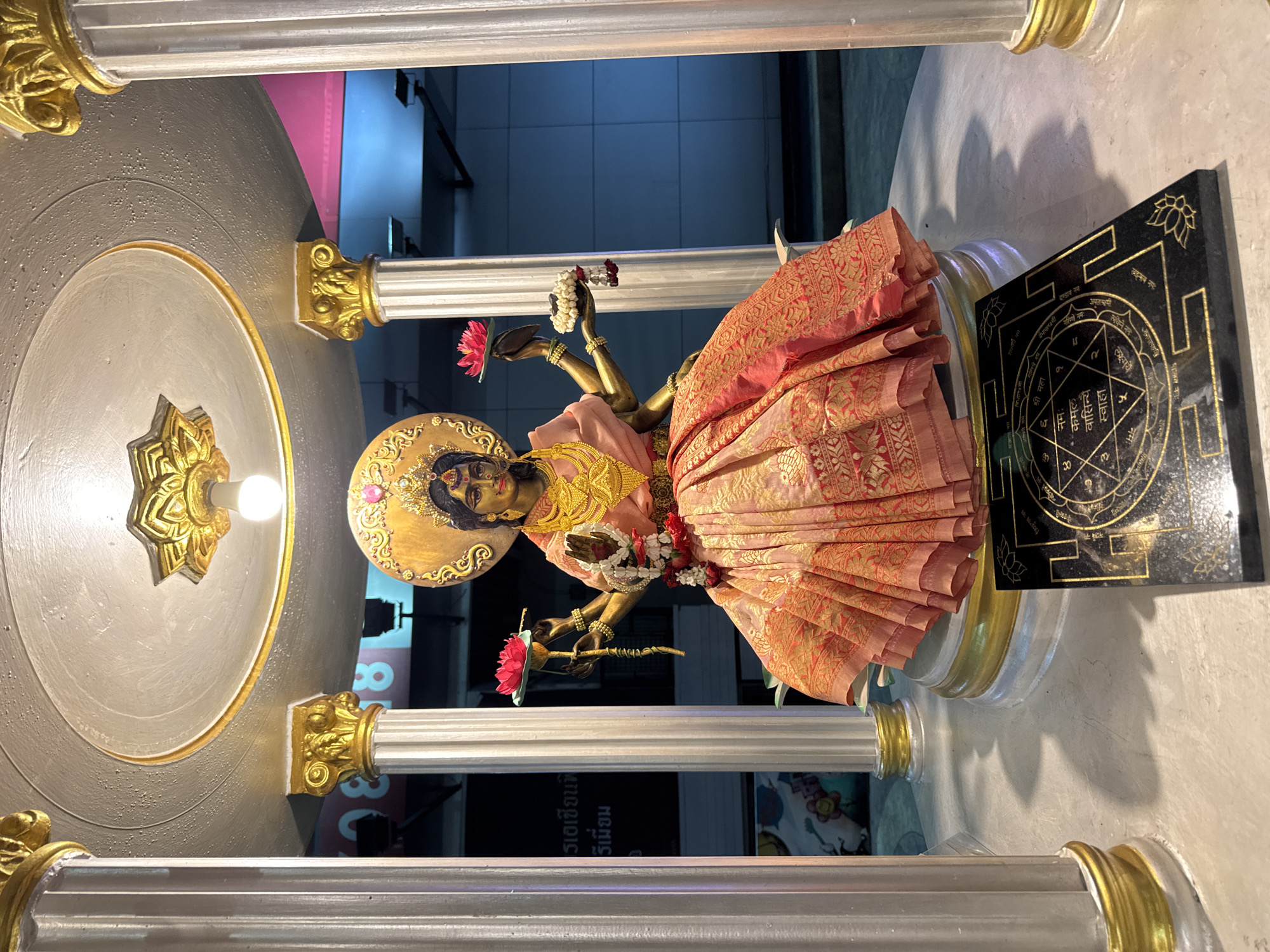 The image shows a statue of Lakshmi, the Hindu goddess of wealth, fortune, and prosperity, in Bangkok, Thailand. The statue is made of bronze and is adorned with gold jewelry and a pink sari. It is standing on a pedestal and is surrounded by pillars and a black tablet with gold lettering in Sanskrit. The statue is placed inside a shop. The background shows a tiled wall and some indistinct objects. The picture seems to be taken indoors.