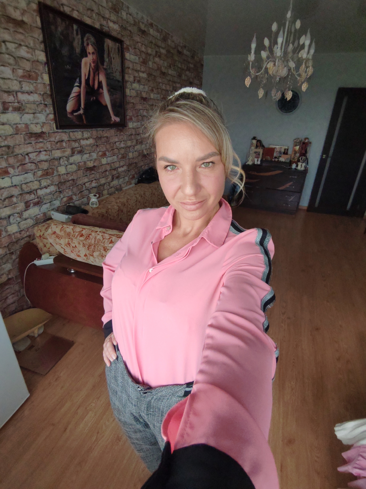 A woman with blonde hair tied in a ponytail and green eyes is taking a selfie in a room. She is wearing a pink collared shirt with black and silver stripes on the sleeves and grey plaid pants. In the background, there is a brick-patterned wallpaper, a framed picture of a woman on the wall, a couch, a dresser with items on top, a chandelier, and a wooden floor. The scene appears to be an indoor residential setting.