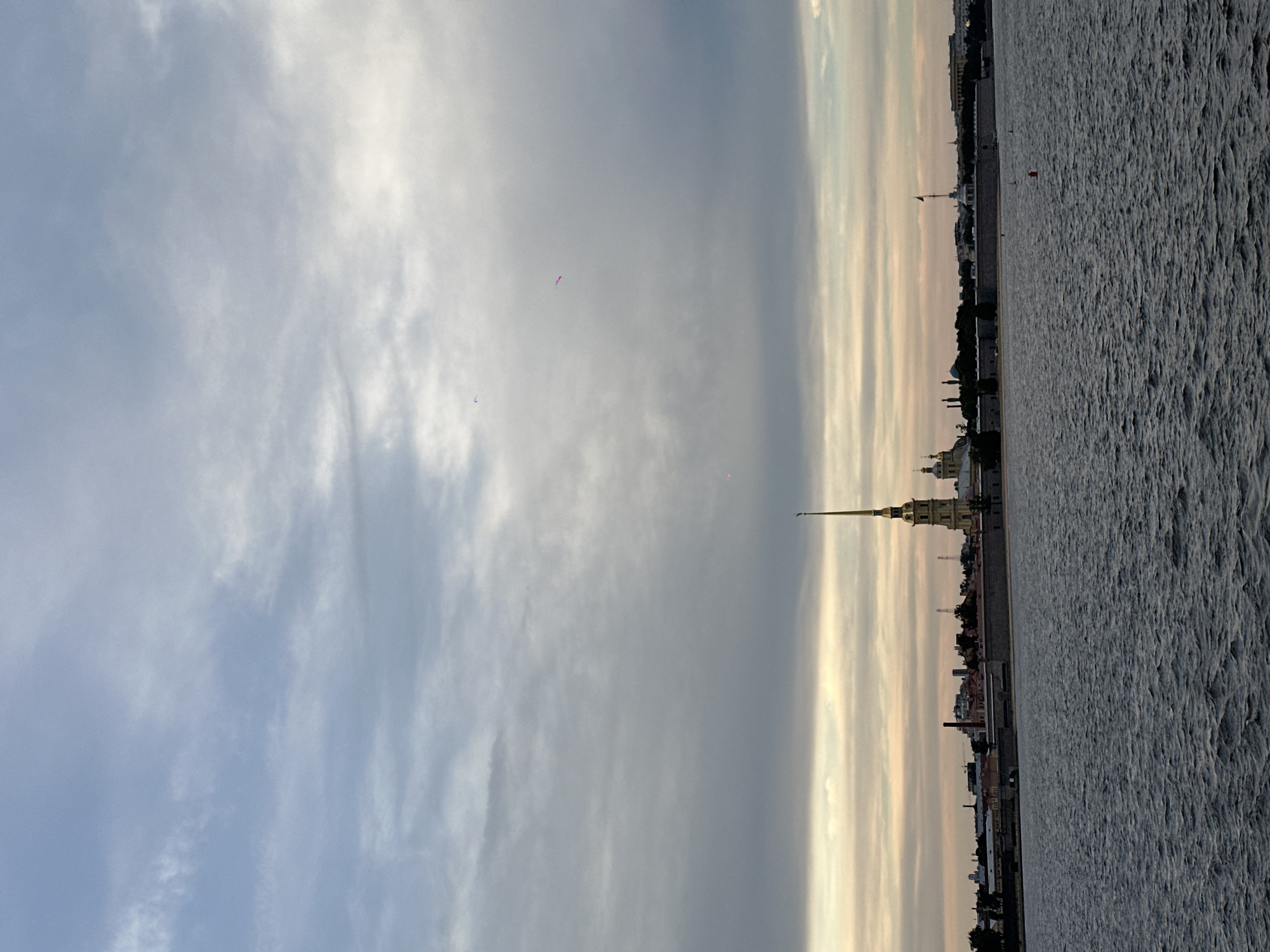 The image shows a wide shot of the Peter and Paul Fortress in Saint Petersburg, Russia, across the Neva River. The sky is overcast with a band of warm, pale yellow light at the horizon, suggesting either sunrise or sunset. The water in the foreground is dark and rippled, reflecting the muted colors of the sky. The fortress complex is visible in the middle ground, dominated by the tall, golden spire of the Peter and Paul Cathedral. The fortress walls are a light gray stone, and behind them, various buildings with domes and chimneys can be seen. A red buoy is visible in the water near the fortress. The overall mood is calm and somewhat melancholic, characteristic of the Saint Petersburg climate.