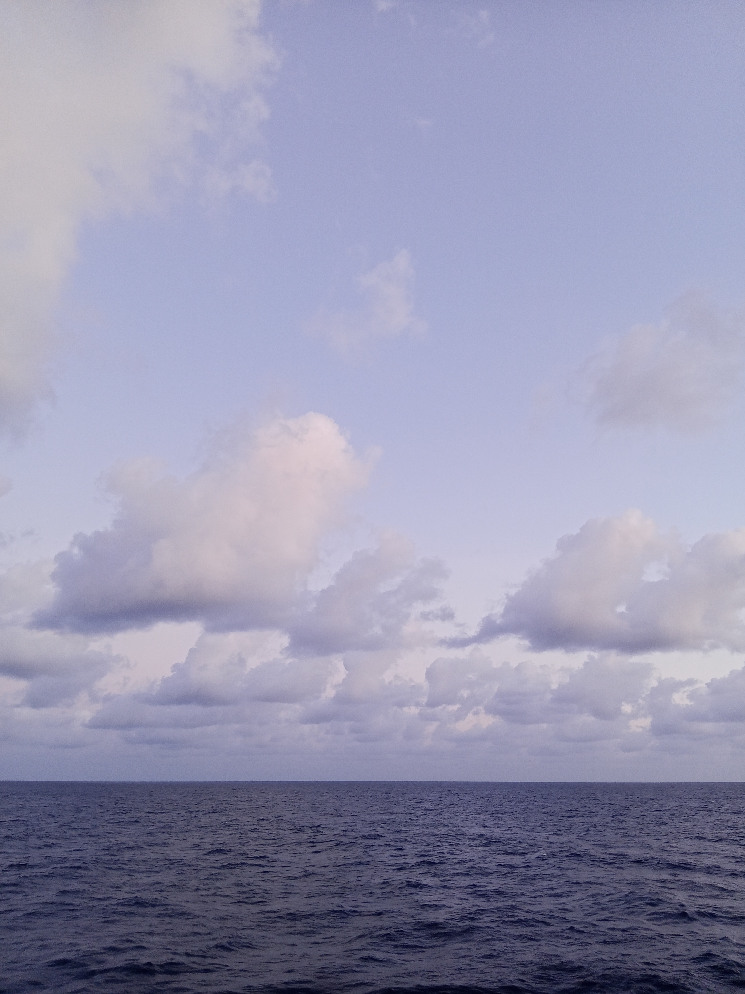 The image depicts a tranquil ocean scene under a pale sky. The horizon is a sharp, straight line separating the dark, undulating water from the sky. The ocean surface is a deep, rich blue, with subtle ripples indicating a gentle breeze or movement. Above, the sky is a soft, muted lavender, filled with scattered clouds of varying sizes and densities. Some clouds are wispy and translucent, while others are more substantial and textured, casting soft shadows. The overall atmosphere is calm and serene, suggesting a peaceful moment at sea. There are no people, buildings, or any other man-made objects visible in the frame. The location is unknown.