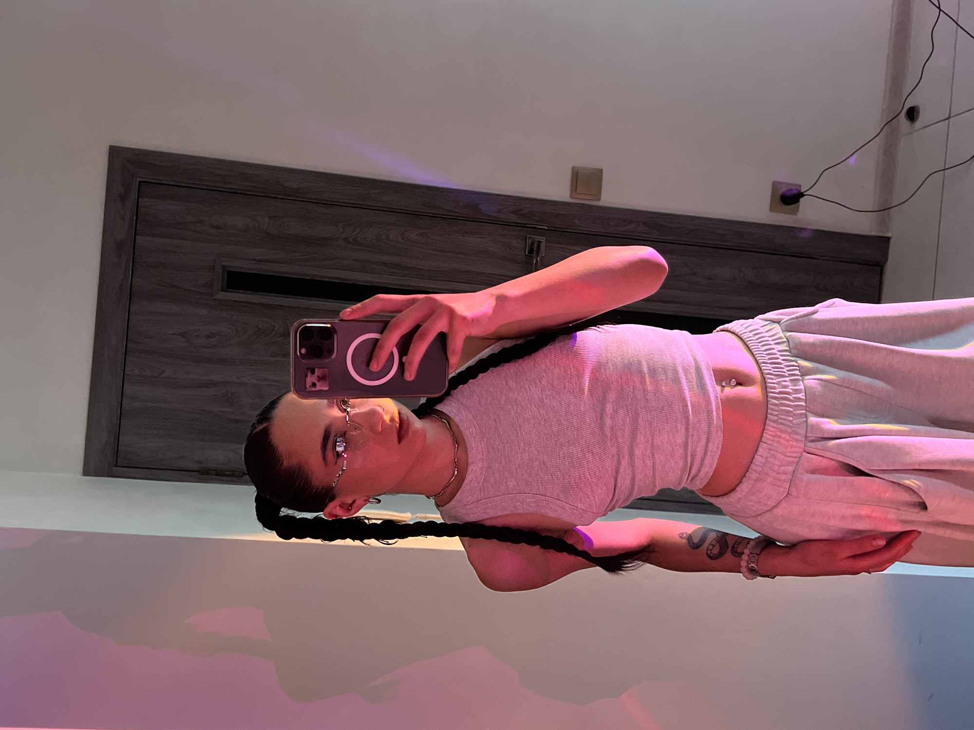 A young woman with dark hair styled in two braids is taking a selfie in a room. She is wearing a grey ribbed tank top, light grey sweatpants, and decorative glasses. A snake tattoo is visible on her left arm. The room appears to be an indoor space, possibly a home or studio, with a grey wooden door in the background and white walls. Colored lights are casting shadows on the wall. There is a power outlet with a cord plugged into it on the right side of the image. The image does not contain any information that can be used to determine its location in Ternopil, Ukraine.