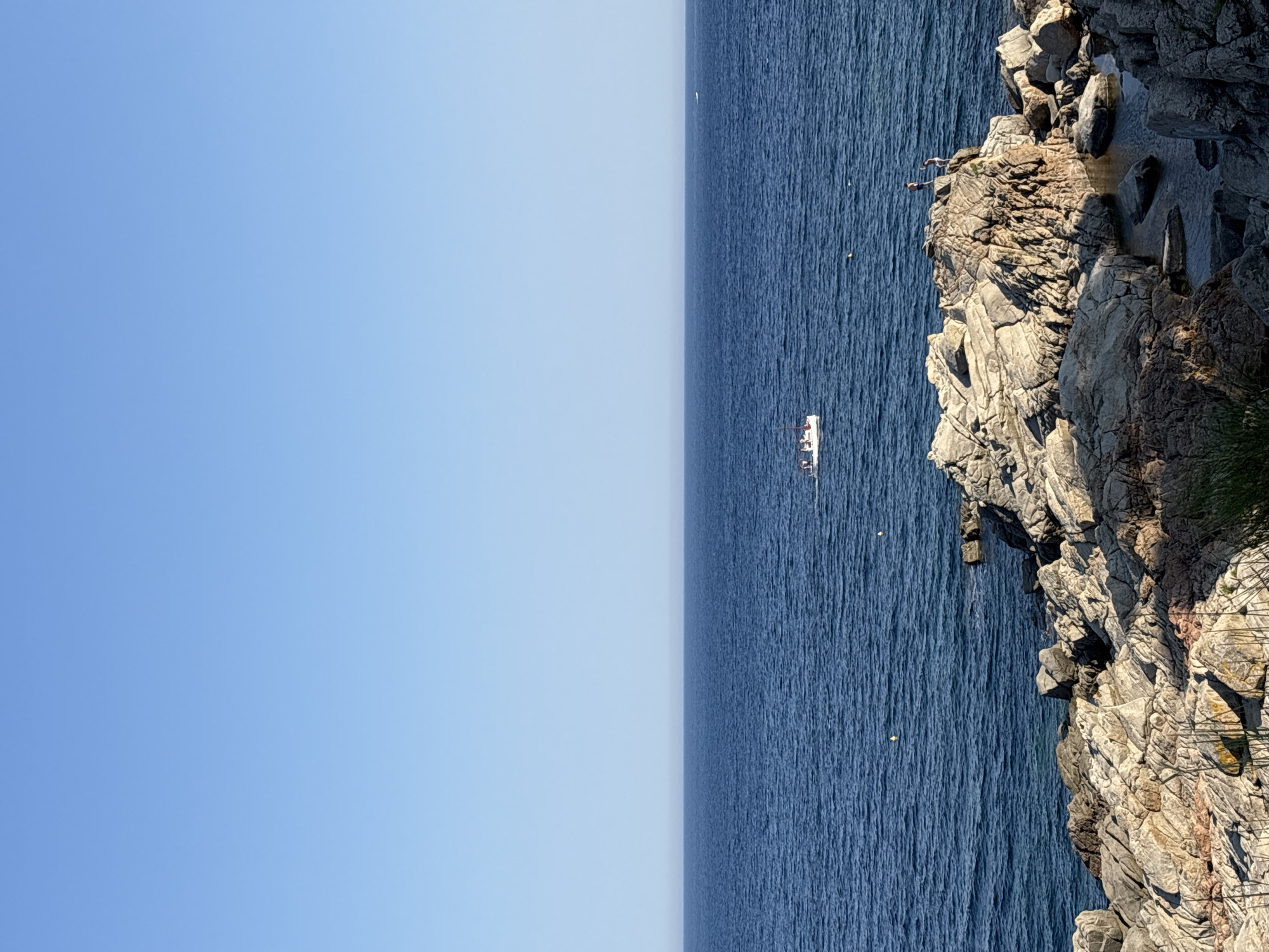 The image captures a serene seascape in Calella De Palafrugell, Spain. The upper portion of the frame is dominated by a clear, expansive blue sky, transitioning seamlessly into the deep blue of the Mediterranean Sea. In the mid-distance, a small white boat with several occupants navigates the calm waters. The foreground is a rugged, rocky coastline, composed of large, weathered grey and tan rocks, interspersed with sparse vegetation. A small inlet of clear water is visible among the rocks. On a prominent rock formation in the foreground, two individuals are standing, appearing to observe the sea or the boat. The overall scene evokes a sense of tranquility and summer leisure.