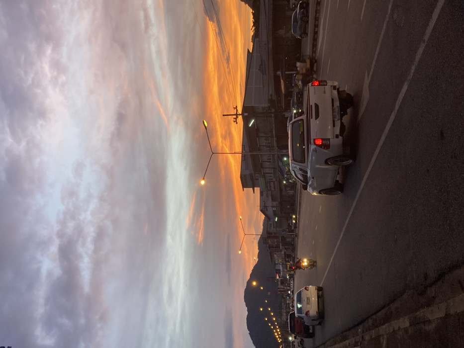 This image captures a multi-lane street scene in Bang Sawan, Thailand, during dusk. The sky presents a vivid sunset, displaying bright orange and yellow hues on the right, gradually fading into purple and grey clouds towards the left and top. Numerous illuminated streetlights, emitting warm yellow light, line both sides of the asphalt road. In the immediate foreground, a white pickup truck, with its red brake lights active, proceeds away from the viewer in the right lane. Further ahead in the left lane, a white sedan and a motorcycle, ridden by a single individual with a lit headlight, are also traveling away from the camera. A red vehicle is visible further down the road on the left. Along the right side of the road, a series of commercial buildings or shophouses, some with illuminated interiors, line the street. Utility poles and power lines are visible above these structures. Distant mountains or hills are silhouetted against the horizon on the far left.