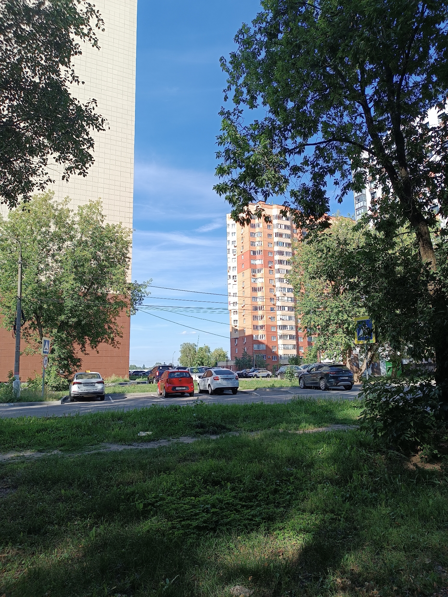 The image captures a street scene in Zhukovsky, Russia, on a clear day with a bright blue sky. In the foreground, lush green grass and trees create a natural boundary. A paved road runs horizontally across the midground, with several cars parked and driving.
Dominating the left side of the frame is a tall, light-colored building with a facade composed of small, square tiles. To its right, a vibrant orange and white high-rise building stands prominently, featuring numerous windows and balconies. Trees are scattered throughout the scene, with some large, leafy trees in the foreground and midground, partially obscuring the buildings and sky.
On the road, a white car with a yellow roof is visible, possibly a taxi, with a license plate that appears to start with "R323TX." A red car and a white car are also present on the road, along with a dark grey SUV. A pedestrian crossing sign with a diagonal pattern and a blue circle containing a white pedestrian symbol is situated near the right edge of the road. A circular traffic sign with the number "20" inside it is attached to a pole on the left side of the road. The overall impression is an urban environment with elements of nature interspersed.