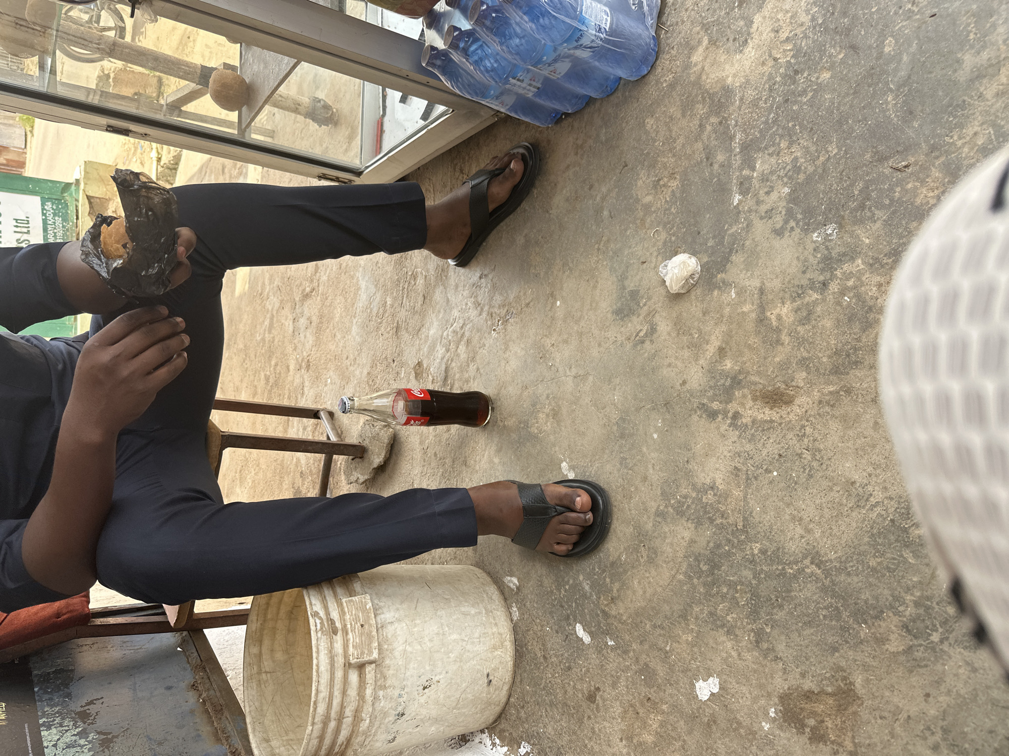 The image depicts a person seated on a chair with their legs crossed, wearing dark trousers and black flip-flops. The person is holding a dark, wrapped food item in their right hand. In front of the person, on the ground, is a bottle of Coca-Cola. To the left of the person is a large, off-white bucket. To the right, in the background, a glass display case contains bottles of water. The scene appears to be outdoors, with a rough concrete ground. The background suggests an urban setting with buildings and a road. A poster with text is visible on the left side of the image. The overall impression is of a casual moment at a roadside vendor or shop in Narayi, Nigeria.