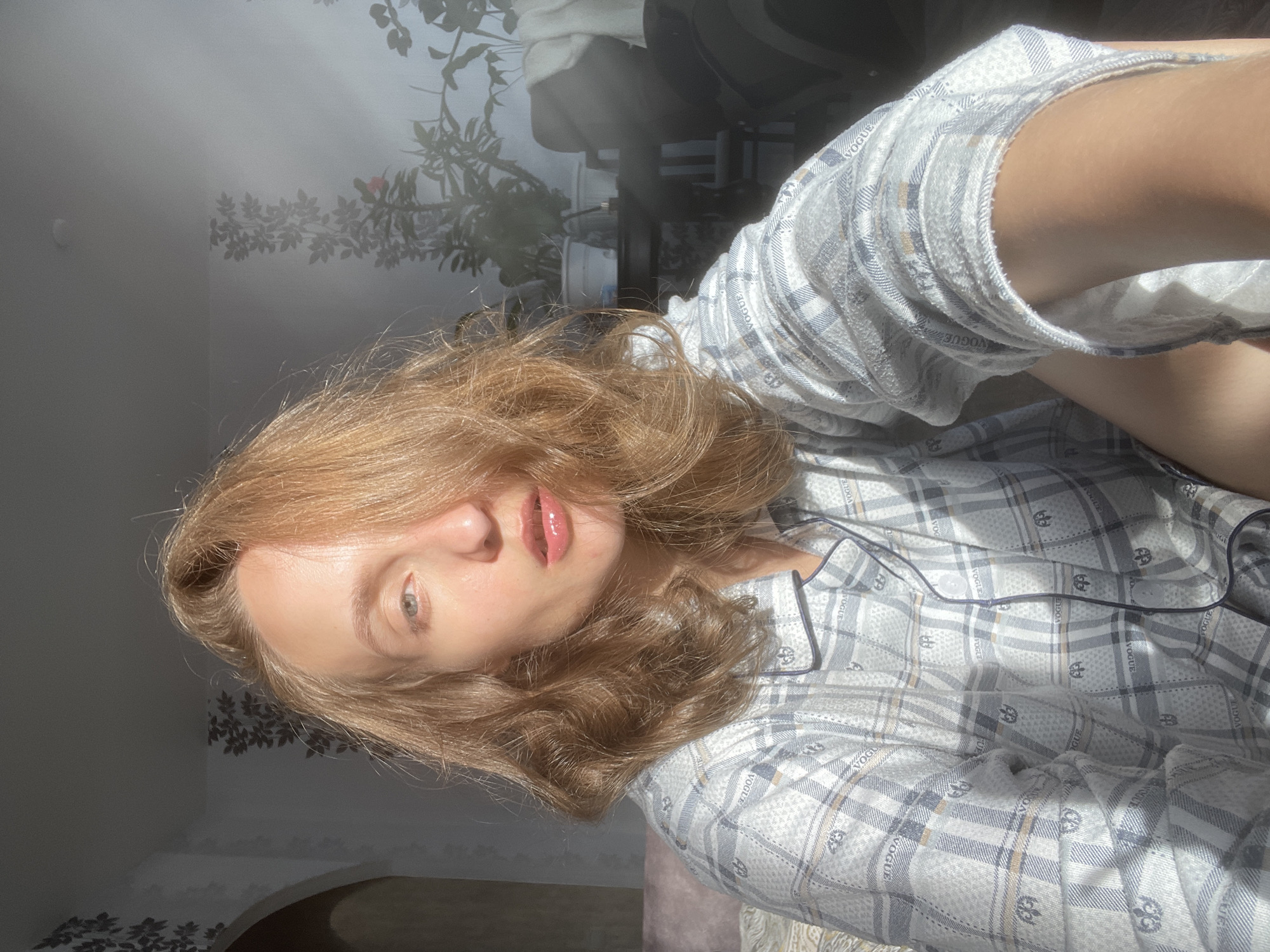 A young Caucasian woman with shoulder-length auburn hair, her face partially obscured by her hair, is seated indoors, likely in a bedroom, as suggested by the soft furnishings and wall decor, possibly in Sharkan, Russia. She is wearing light-colored pajamas with a plaid pattern and subtle branding. The sunlight streams in from the right, casting dramatic shadows and highlighting her features. The background reveals a wall with decorative wallpaper featuring a floral motif and a glimpse of plants in pots, indicating an indoor residential setting. The overall impression is a candid, natural moment captured in the morning light.