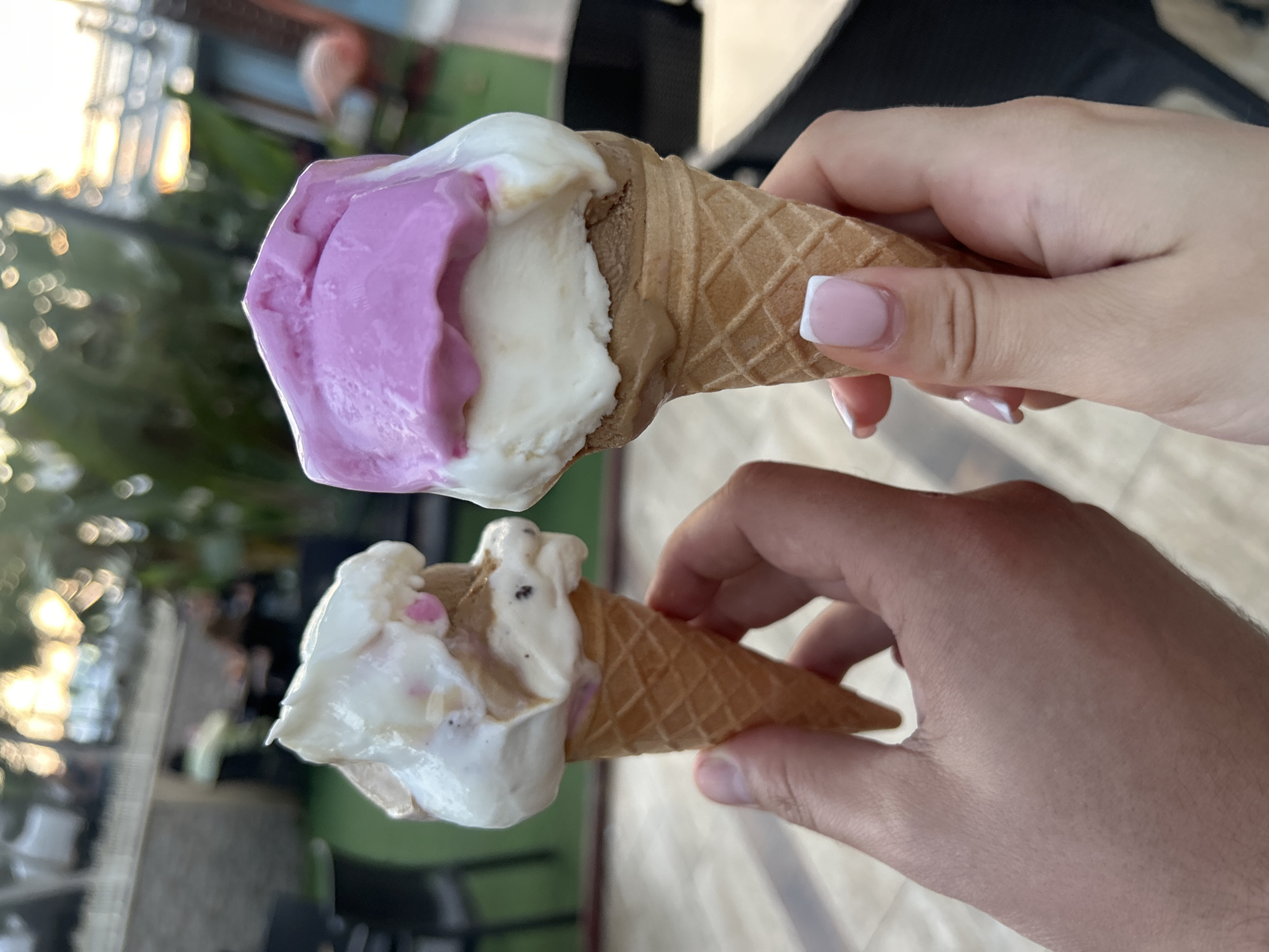 Two hands are holding ice cream cones outdoors. The top cone is held by a hand with pink and white manicured nails and contains scoops of purple, white, and brown ice cream. The bottom cone is held by a male hand and contains scoops of white and brown ice cream. The location appears to be a city, as suggested by blurred buildings and foliage in the background. The city is Unknown, Unknown.
