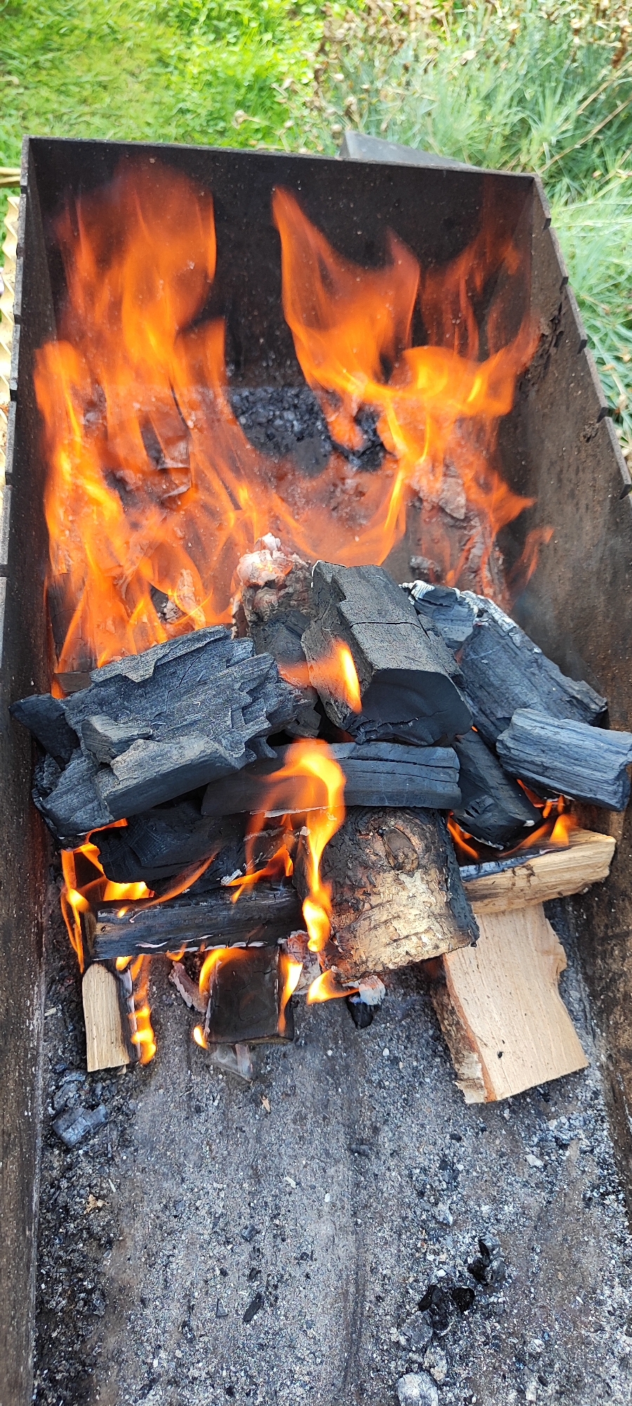 This is a high-angle, close-up shot of a barbecue grill filled with burning charcoal and wood. The flames are a vibrant orange and yellow, consuming the fuel and casting a warm glow. Large pieces of charcoal, some partially ignited, are scattered throughout the grill. Smaller pieces of wood, including a few with bark still attached, are also visible, some catching fire. The bottom of the grill is covered in ash and embers. The background shows a blurry expanse of green grass, suggesting an outdoor setting, possibly in the rural area of Лёзненскі сельскі Савет, Belarus. There are no people or animals present in the image, and the focus is solely on the fire and the grilling apparatus.