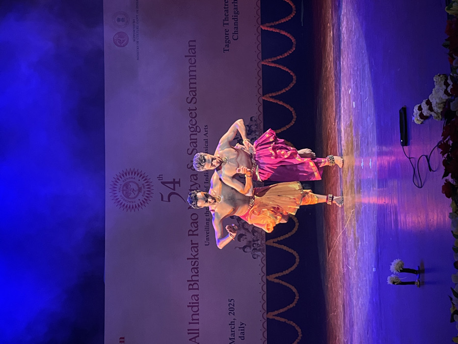 The image shows two men performing a dance on a stage in the Tagore Theatre in Chandigarh, India. The stage is lit with purple and blue light, and the floor is made of polished wood. The men are wearing traditional Indian clothing: one is wearing a gold and orange outfit and the other is wearing a pink and gold outfit. The background is a large, white wall with the text "All India Bhaskar Rao Natya & Sangeet Sammelan" in large letters, along with the text "54th", "Unveiling the Soul of Indian Classical Arts", "March, 2025 daily", and "Tagore Theatre Chandigarh". There is a garland of flowers strung along the wall, as well as a small microphone and a black cord lying on the stage floor. The image was taken at coordinates (latitude: 30.733, longitude: 76.789).