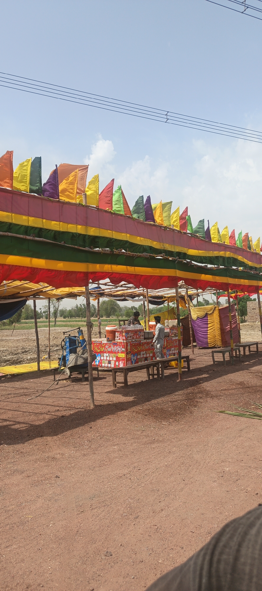 This image captures a vibrant outdoor refreshment stall in a rural or semi-rural setting in Mandihar, Pakistan, under a clear daytime sky. The central feature is a long, temporary canopy constructed from multiple layers of brightly colored fabric in red, yellow, green, and pink stripes, supported by wooden poles. Along the top of the canopy, numerous small, colorful flags – orange, yellow, green, purple, and red – flutter in the breeze, creating a festive atmosphere.
Underneath the canopy, a stall or counter is visible, decorated with colorful patterns and text, primarily red. Two men are present: one stands behind the counter, appearing to be attending to the stall, while another is partially visible. On the counter, two orange and white insulated containers are placed, likely holding beverages. To the left of the stall, a blue and white machine, possibly a sugarcane juice extractor or water pump, is covered with fabric. Two simple wooden benches are positioned in front of the stall for customers.
The ground is unpaved, consisting of reddish-brown dirt, with shadows indicating bright sunlight. In the background, beyond the structures, are open fields, some appearing cultivated or fallow, leading to a distant line of trees. Overhead, several power lines stretch across the expanse of a light blue sky dotted with scattered white clouds.
Visible Urdu text on the stall prominently displays "ٹھنڈا اور تازہ شکنجبین" (Thanda aur taza shikanjbeen), translating to "Cold and fresh lemonade/shikanjbeen," indicating the primary product sold. Other text appears to include "بچہ بازار" or "بچوں کا بازار," suggesting a "children's market" or general small market. The scene depicts a functional, community-oriented refreshment point, likely popular in warm weather.