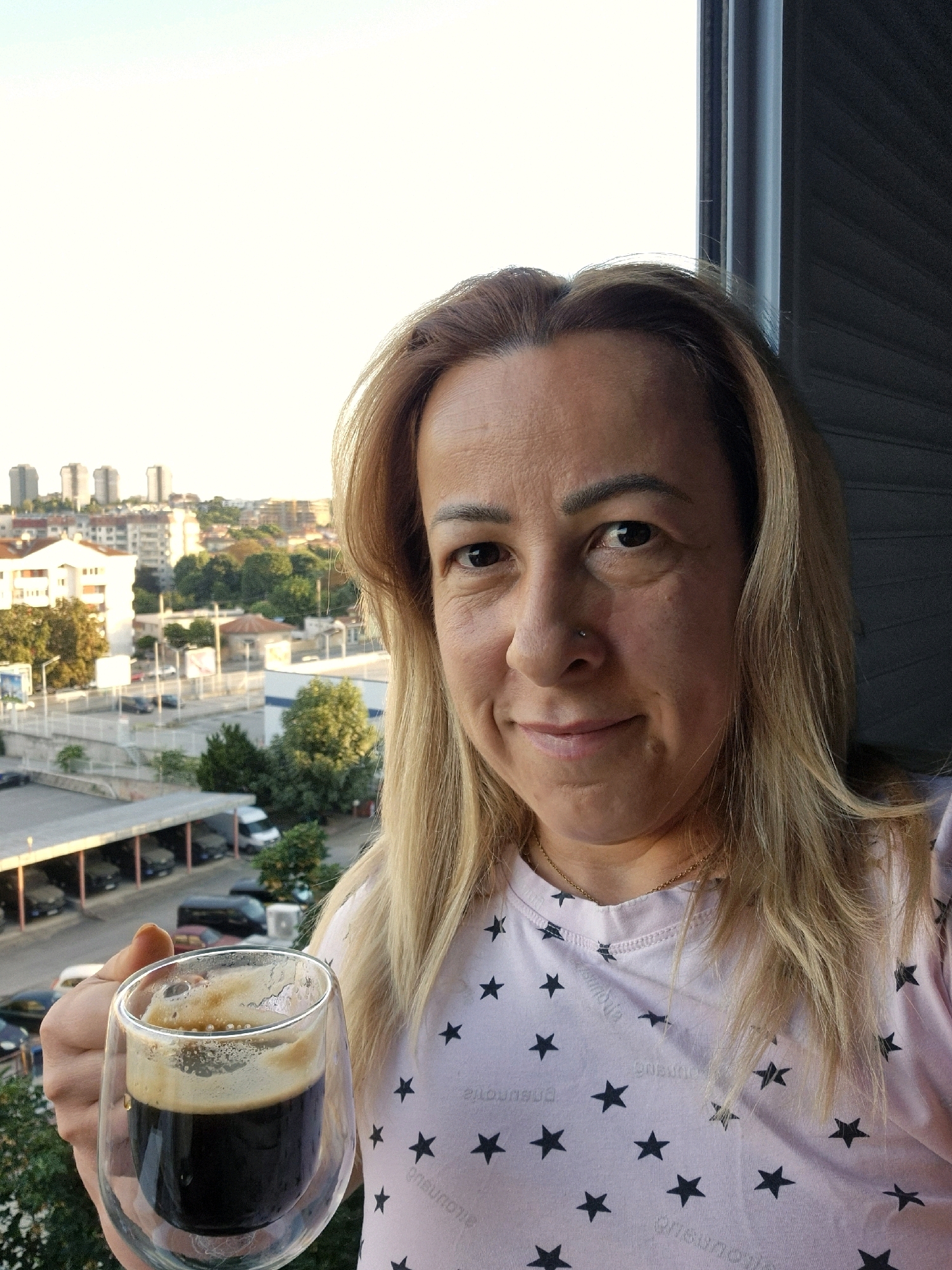 A woman with blonde hair is holding a glass of coffee in her left hand. She is wearing a pink t-shirt with black stars on it. In the background, there is a city skyline with buildings and trees. The sun appears to be setting, casting a warm glow over the scene.