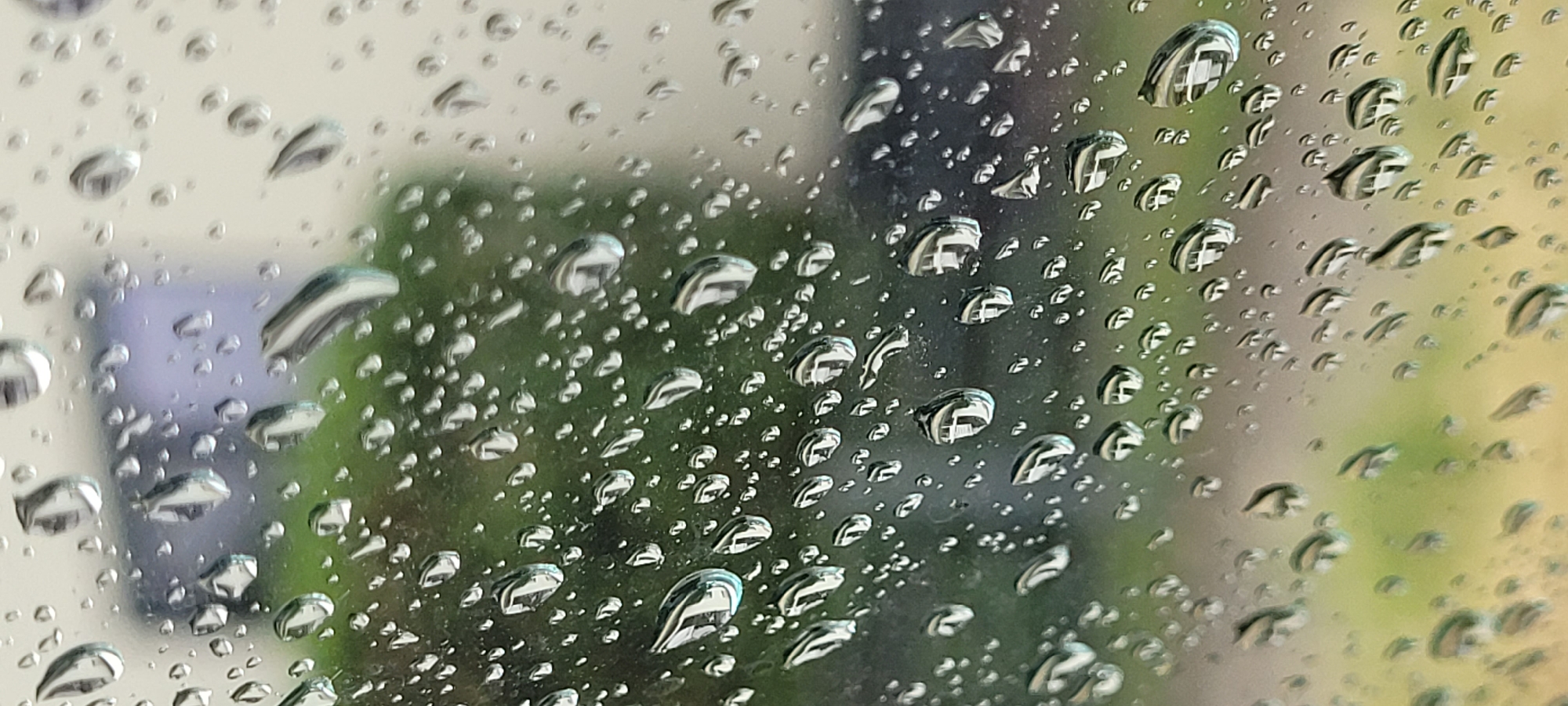 The image shows a close-up view of raindrops on a window, with a blurred background of greenery and a light-colored building, possibly in Dietzenbach, Germany. The raindrops are varied in size and shape, some appearing as perfect spheres while others are elongated or distorted. They refract and reflect the light and the surrounding environment, creating interesting visual effects. The green foliage in the background suggests an outdoor setting, perhaps a garden or park, while the blurred building indicates urban or suburban surroundings. The overall impression is one of a rainy day, with the window acting as a barrier between the viewer and the outside world.