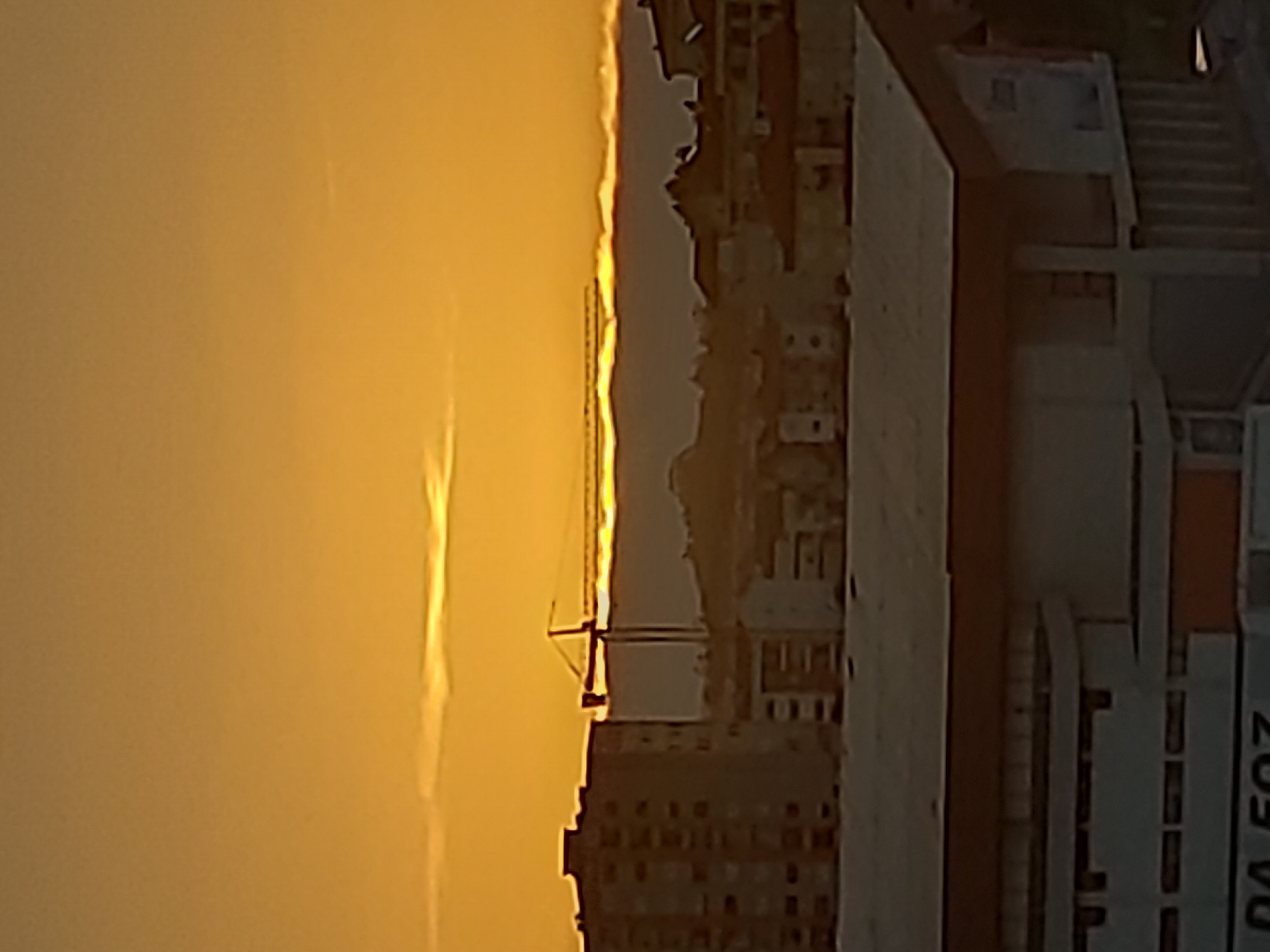 The image shows a view in Figueira da Foz, Portugal. In the foreground, part of a building with the text "DA FOZ" is visible. Beyond the building is a cityscape fading into a golden sky, likely during sunset or sunrise. A crane stands out against the bright sky near the buildings.