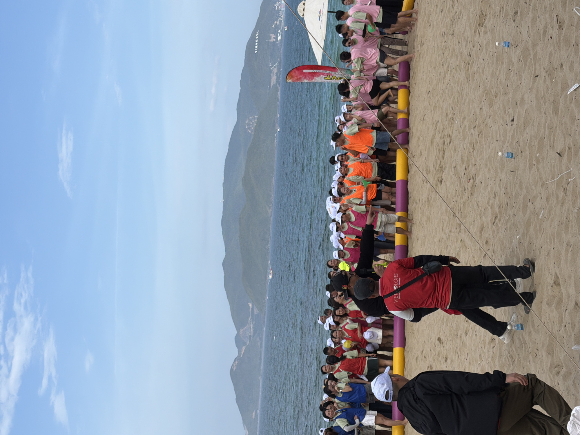 The image depicts a group of people on a sandy beach in Nha Trang, Vietnam, with the ocean and a mountainous coastline in the background. The sky is partly cloudy and a boat is visible in the distance on the water. The people, many wearing similar colored vests (pink and orange) and some wearing red shirts, appear to be participating in an outdoor event or team-building activity. There is a barrier made of large colorful tubes in the foreground and some bottles are placed on the sand. The overall atmosphere seems to be a casual, outdoor gathering. The Vinpearl logo is visible on the hillside in the distance.
