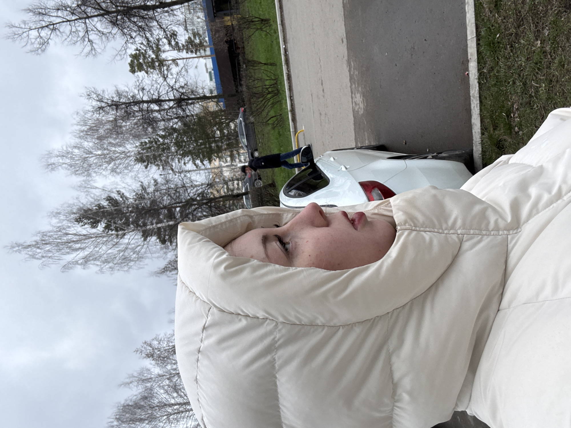 A portrait-oriented outdoor photograph features a young person, likely female, prominently in the foreground, wearing a light-colored (cream or beige) hooded puffer jacket. Her face is shown in profile, looking upwards with a neutral, contemplative expression. The jacket's hood is up, framing her face.
The background depicts an urban or suburban environment under an overcast sky, suggesting daytime in a cool season. Bare deciduous trees with intricate branches dominate the upper and left sections of the frame, indicating late autumn, winter, or early spring. Some evergreen trees are also visible.
An asphalt road or paved area runs horizontally across the mid-ground, marked by a white line. Patches of grass and sparse vegetation are present along the roadside. In the distance, faint architectural structures, including a building with a blue roof, can be seen. A small, indistinct figure is visible further back on the road, appearing to ride an electric scooter or bicycle. A white car is parked on the roadside, partially visible behind the main subject, showing its rear section and a red taillight.
The scene conveys a calm, outdoor moment. No visible text is present in the image.