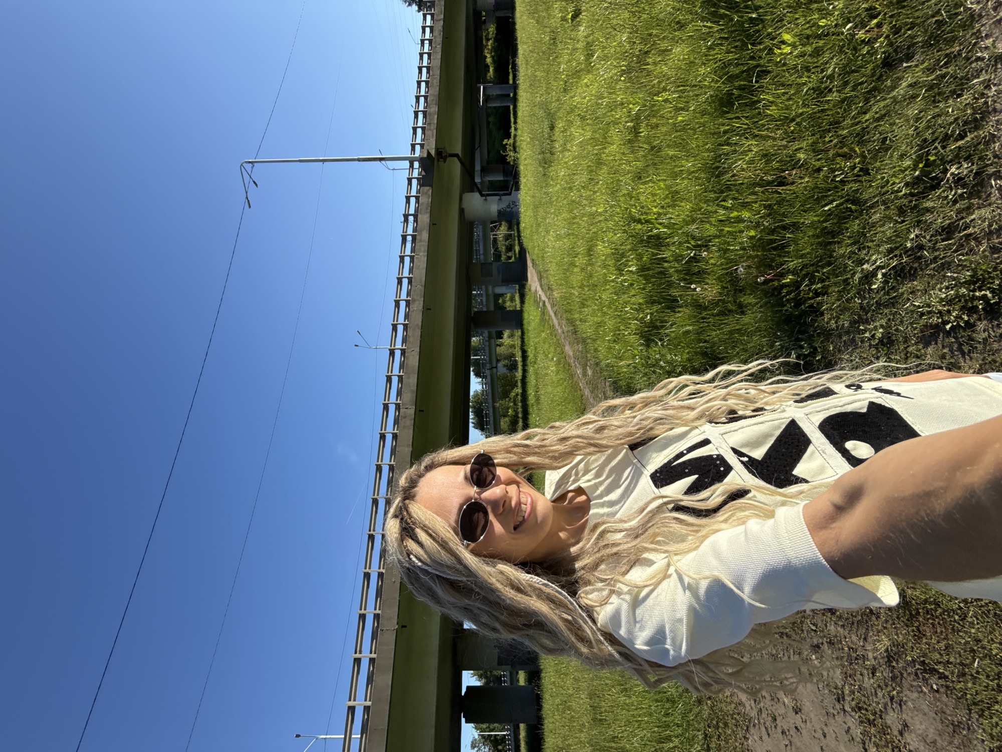 An outdoor selfie captures a light-skinned female with long, blonde dreadlock-style hair, wearing dark, oval-framed sunglasses and a light-colored, short-sleeved top with large black patterns, possibly sequins. She is smiling, revealing her teeth, and extends her right arm to hold the camera. The setting is a grassy field with a narrow dirt path, under a clear blue sky. In the background, a large concrete overpass or bridge structure with multiple support pillars and a metal railing spans the scene. Overhead utility wires and lampposts are visible against the sky and near the bridge. This scene is located in the city of Шебунино, Russia, under bright daylight conditions.