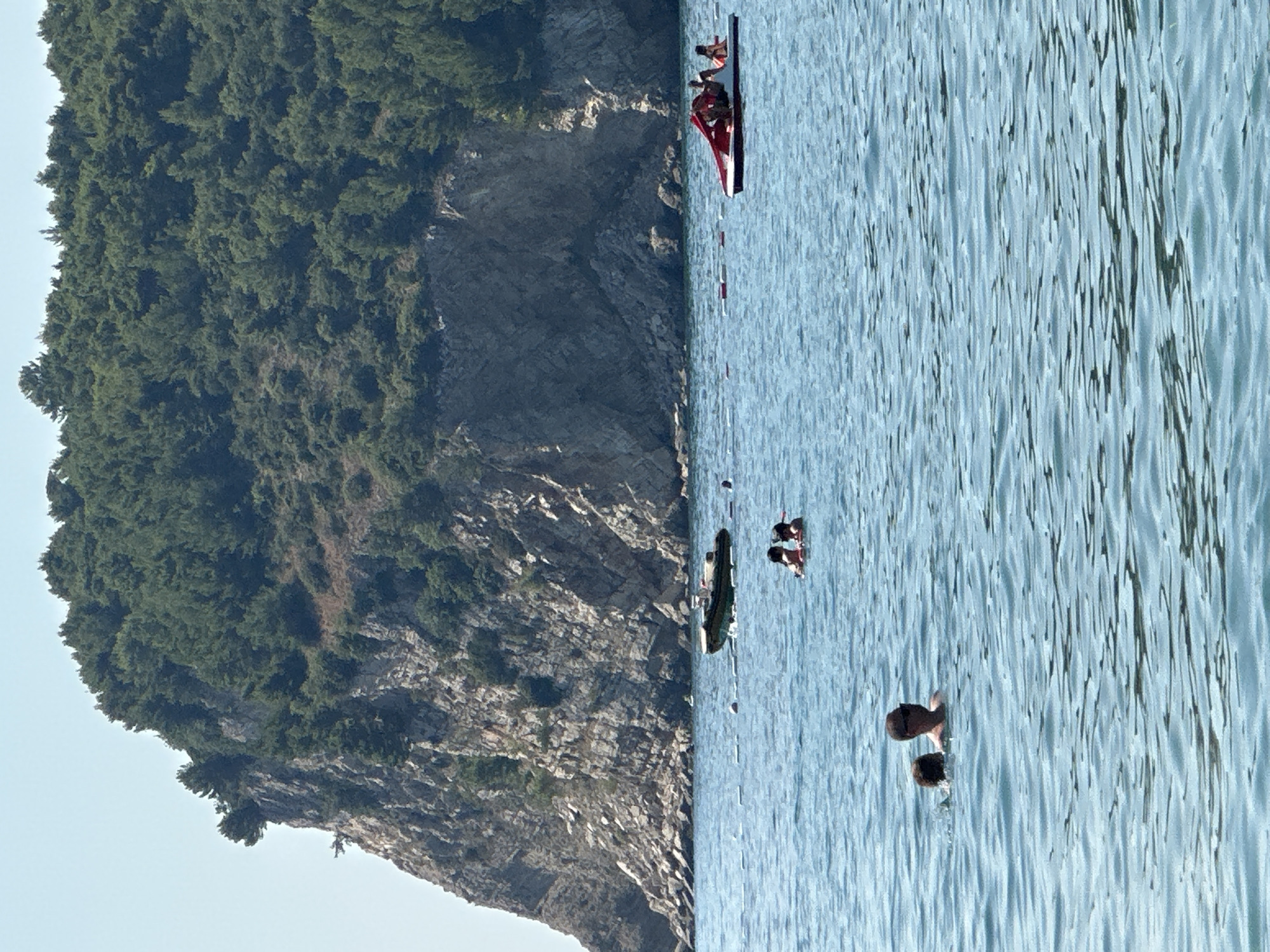 In Đurovići, Montenegro, an image depicts a serene seascape with a prominent rocky cliff face covered in dense green foliage dominating the background. The calm, blue water in the foreground is dotted with several individuals and watercraft. In the middle ground, a small, inflatable boat is anchored, and further to the right, a red and white paddle boat carries several passengers. Closer to the viewer, two people are visible in the water. One appears to be a man wearing sunglasses and a dark shirt, with a second person partially submerged beside him. A cluster of people is also present further out in the water, near the anchored boat. The sky is a clear, pale blue.