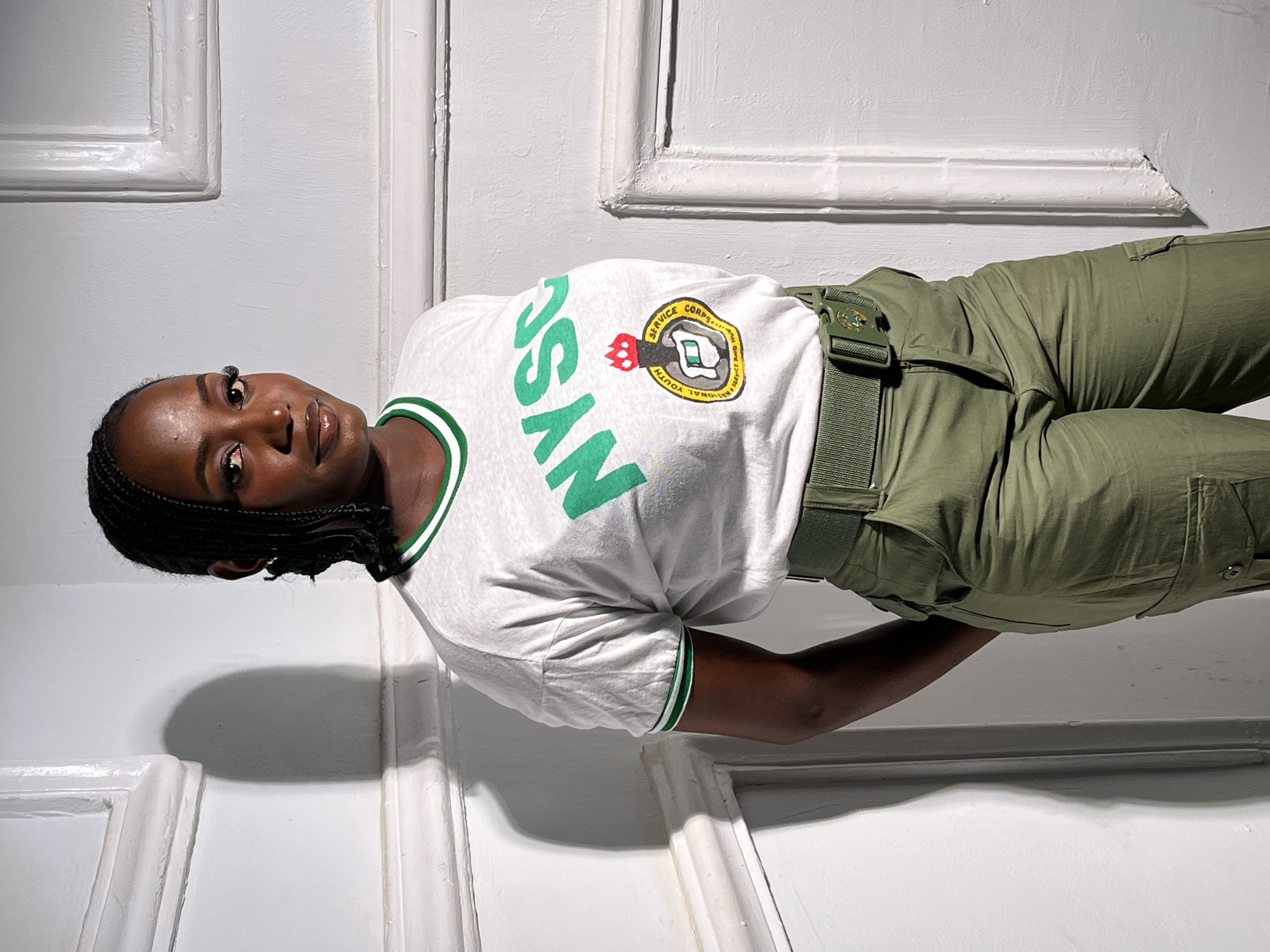 The image depicts a woman wearing the uniform of the National Youth Service Corps (NYSC) of Nigeria. She is wearing a white t-shirt with green trim on the sleeves and collar, printed with the NYSC logo and acronym "NYSC" in green letters. She is also wearing green khaki trousers and a green belt. The woman has dark skin and braided hair. She is positioned at an angle to the camera, with a white wall and door frames visible in the background. The location in the city of Jos, Nigeria, is not discernible from the image.