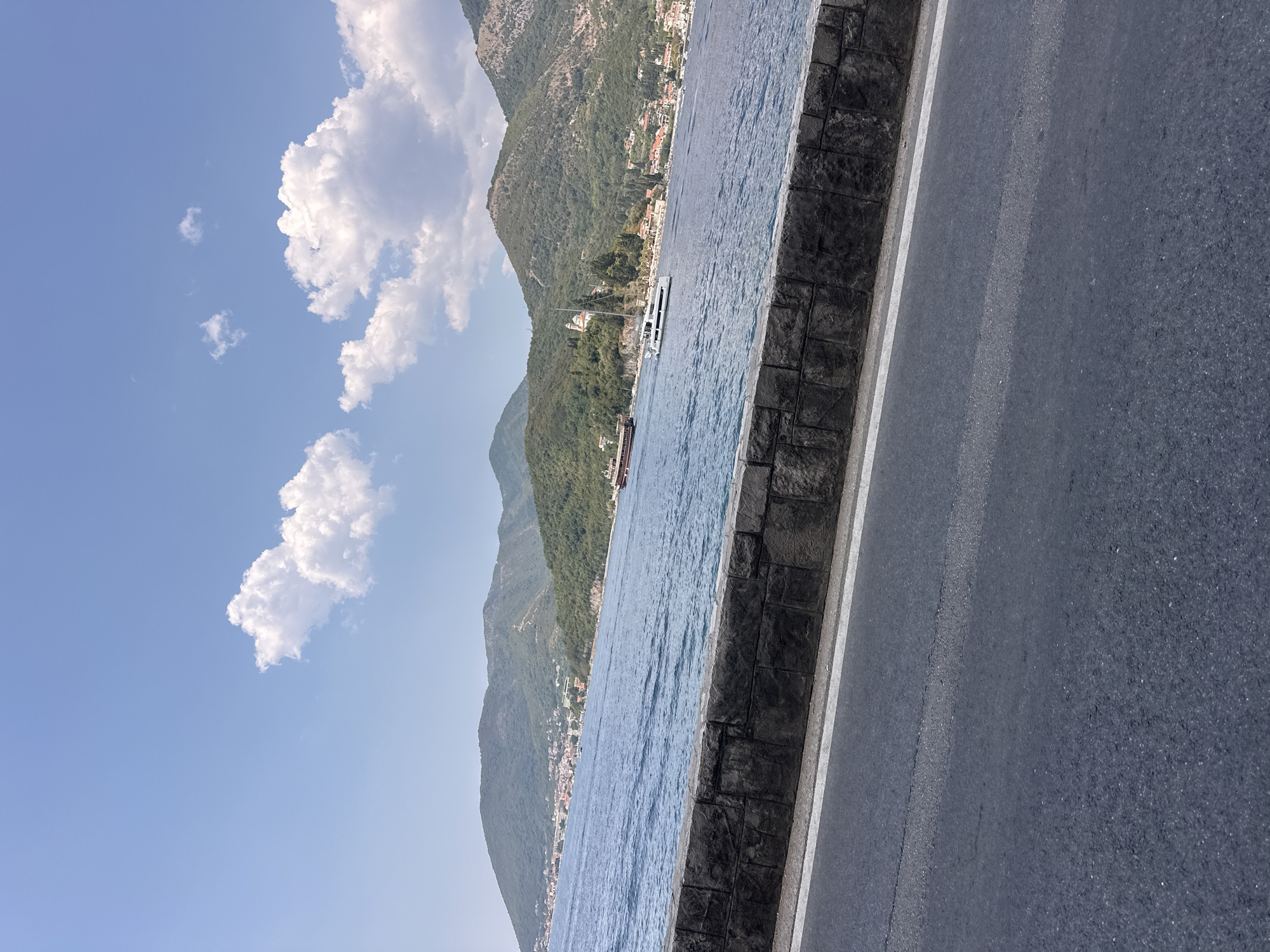 The image depicts a scenic view of the Bay of Kotor from the town of Lepetane, Montenegro. In the foreground, a dark asphalt road with a white line runs parallel to a textured stone wall. Beyond the wall, the deep blue water of the bay stretches out, reflecting the clear sky. Several boats are visible on the water, including a large red and white vessel and a sleek white yacht. The background is dominated by lush green, tree-covered mountains that rise steeply from the water's edge. A small town with red-tiled roofs is nestled along the shoreline, with buildings clinging to the slopes of the hills. The sky is a vibrant blue, dotted with fluffy white clouds. The overall scene conveys a sense of tranquility and natural beauty.