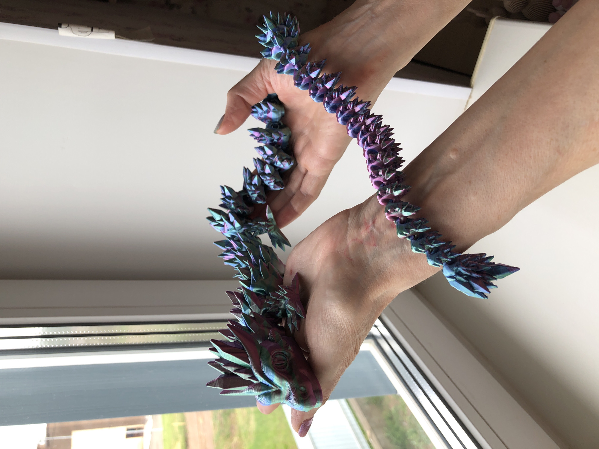 A person is holding a multi-colored, articulated dragon figure in their hands. The figure is primarily blue and purple with a scale-like texture. The hands are Caucasian and show signs of aging, such as wrinkles and slight redness. The background includes a window with a view of an exterior landscape with grass and shrubbery, as well as a white wall. The image does not contain location information about Noviy, Russia.