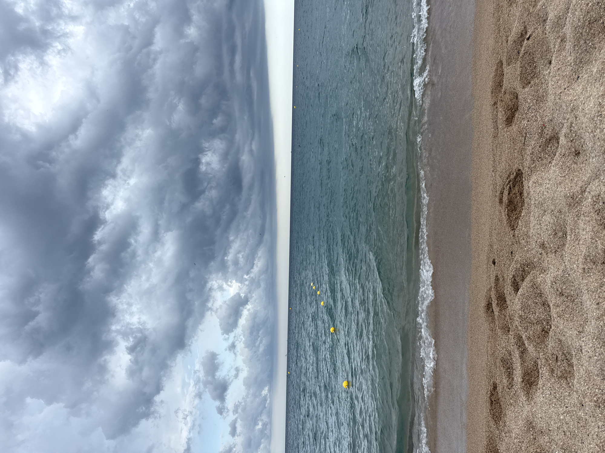 A beach scene in Blanes, Spain, is depicted under a cloudy sky. The foreground is a sandy beach with footprints. The middle ground features the turquoise ocean with small waves lapping at the shore. A line of yellow buoys stretches across the water. The background is dominated by a horizon of dark, heavy clouds, suggesting an approaching storm. The sky is a mixture of grays and blues, with some lighter patches.