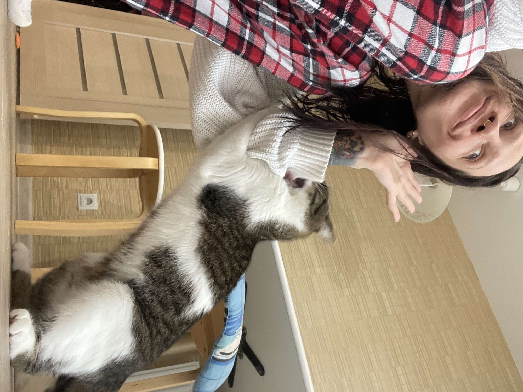 A woman with a nose piercing and tattoos on her arm is holding a gray and white tabby cat. She is wearing a white sweater and a red and black plaid shirt. They are in a room with wooden furniture and patterned wallpaper. The location is Izhevsk, Russia.