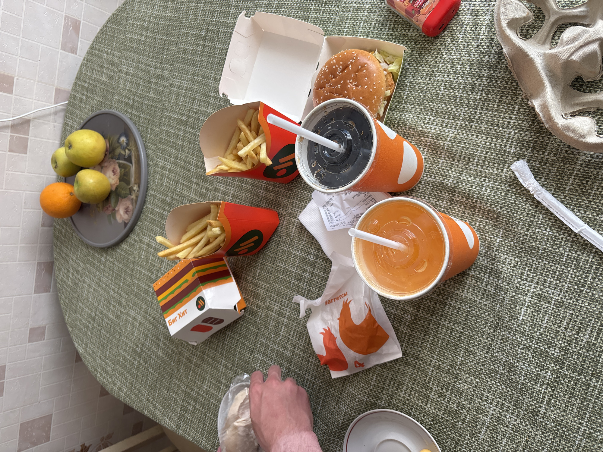 A bird's-eye view captures a table setting for a meal, likely a takeout order, on a green textured tablecloth. The scene includes two orange paper cups with straws, one filled with a dark liquid and the other with an orange beverage. Adjacent to the cups are two red and orange french fry containers, both filled with golden fries. A burger in a white paper box sits next to one of the cups. A crumpled orange and white paper wrapper with a shrimp graphic is visible in the foreground. To the left, a hand in the process of holding a plastic bag of food is partially visible. In the upper background, a plate holds an orange and three green apples. The room's tiled wall is visible behind the table. A red ketchup bottle and a cardboard tray are also present on the table. The location is identified as Sinyavino, Russia.