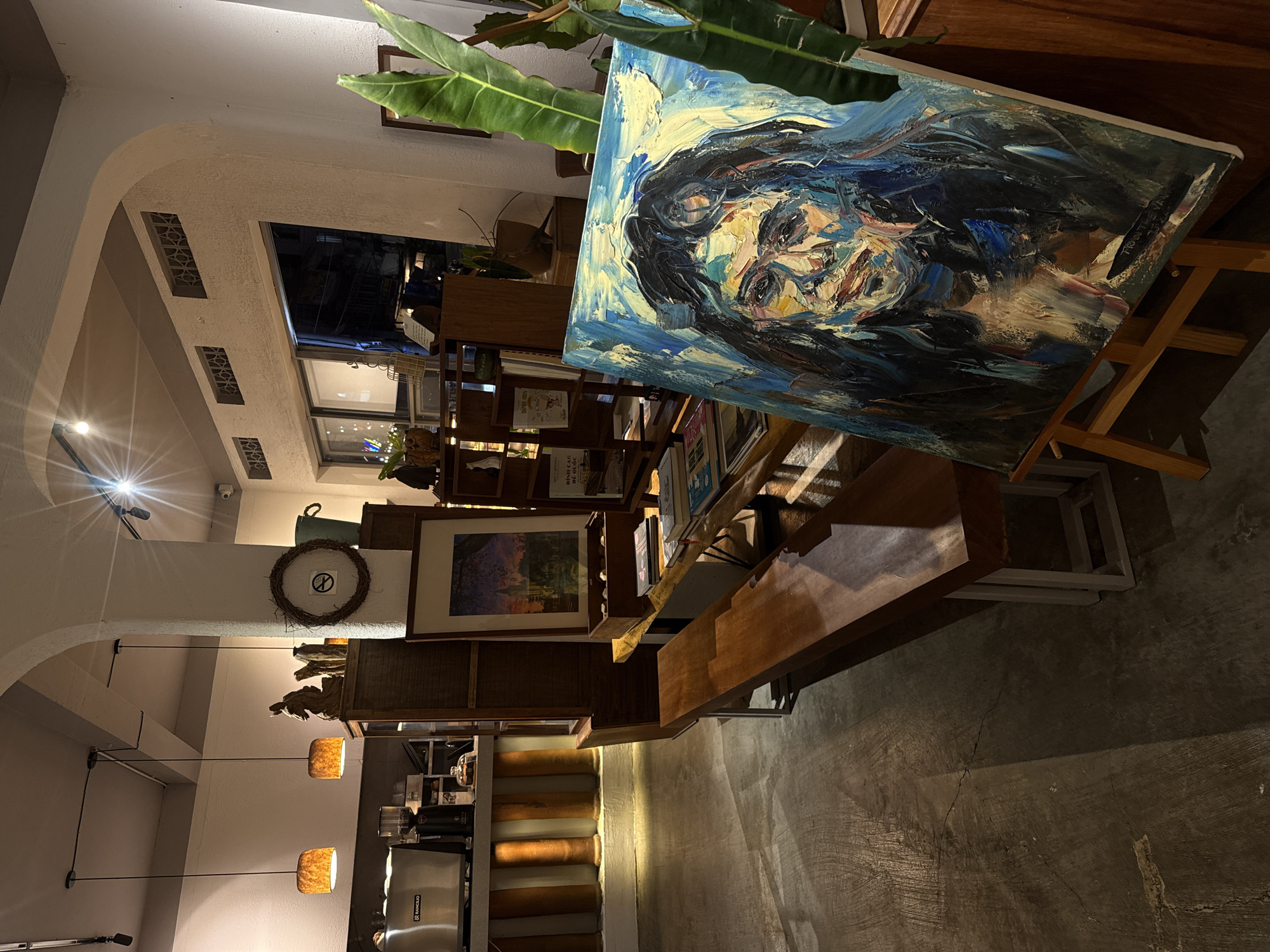 This is a photograph of an art display within what appears to be a cafe or shop in Da Lat, Vietnam. The main subject is a large, impressionistic oil painting of a woman's face, rendered with thick, textured brushstrokes in shades of blue, yellow, and black. The painting is propped on a wooden easel.
The setting is indoors, with warm, ambient lighting casting soft shadows. In the background, there are shelves filled with books and decorative items, including a framed landscape painting and a wreath with a "no smoking" symbol. The architecture features a curved archway and ventilation grates. Hanging pendant lamps with warm light illuminate the space. The floor is a polished concrete surface.
The time of day is likely evening or night, given the indoor lighting and the darkness visible through a window in the background, which hints at the outside environment. The overall mood is calm and artistic, suggesting a space dedicated to creativity and relaxation.
Visible text includes "TRANTHE VAN" inscribed on the right edge of the painting. Some Vietnamese text is also visible on book spines and potentially other displayed items, though it is largely illegible in this image.