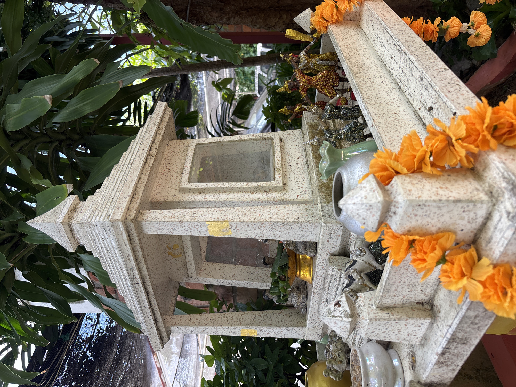 The image depicts a spirit house, likely located in Chalong, Thailand, featuring a multi-tiered structure made of speckled white material, possibly concrete or stone. The house is adorned with ornate carvings and has several small windows. Within the structure, there are small figurines, some appearing to be human-like figures in traditional attire, and a golden offering bowl. The exterior of the spirit house is decorated with garlands of bright orange flowers. In the foreground, there are more decorative elements, including small elephant statues and a white ceramic bowl filled with pebbles. The spirit house is situated outdoors, surrounded by lush green foliage and trees, with glimpses of brickwork and possibly a building in the background. The overall scene suggests a place of worship or offering, common in Thai culture.