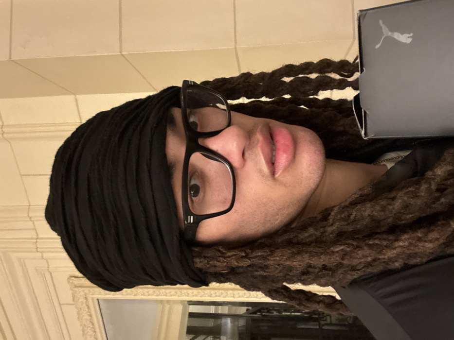 An individual with medium-brown skin, wearing a black wrapped head covering and black-rimmed rectangular glasses, is depicted in a close-up indoor shot. Long, dark brown dreadlocks are visible, framing the face and extending over the shoulders. The individual wears a dark-colored top and a silver chain around the neck, and exhibits a light stubble. A dark grey or black box with a visible white or silver leaping cat logo, consistent with the Puma brand, is held in the lower right portion of the frame. The background features light-colored, ornate architectural details: a decorative mirror or panel with an intricate frame on the left, reflecting a dark interior, and a cream-colored wall with vertical pilasters or columns on the right. The setting suggests a formal interior space, such as a hotel lobby, historic building, or upscale residence within Paris, France, though the exact street address is not identifiable.