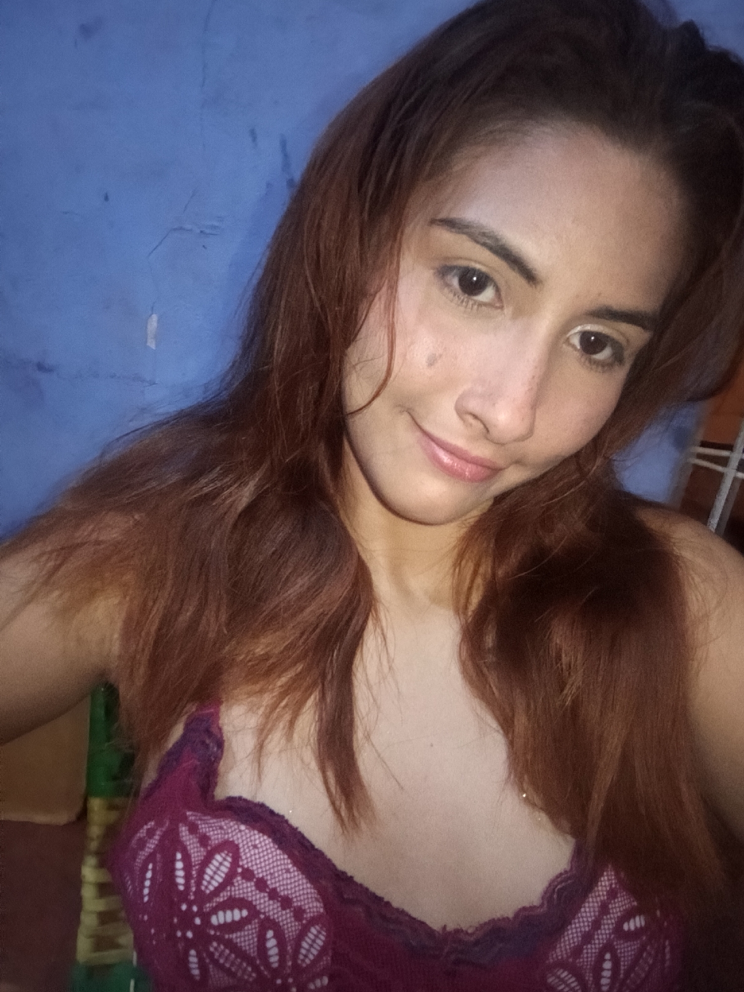 A young woman with reddish-brown hair and light brown eyes is pictured from the chest up, looking directly at the camera. She is wearing a purple lace bra. Her skin has some freckles. The background is a blue wall with some texture and discoloration. To the left of the woman, a green object with some yellow stripes is visible, possibly a piece of furniture. The lighting suggests an indoor setting. The image appears to be a selfie taken in the city of Juan Griego, Venezuela.