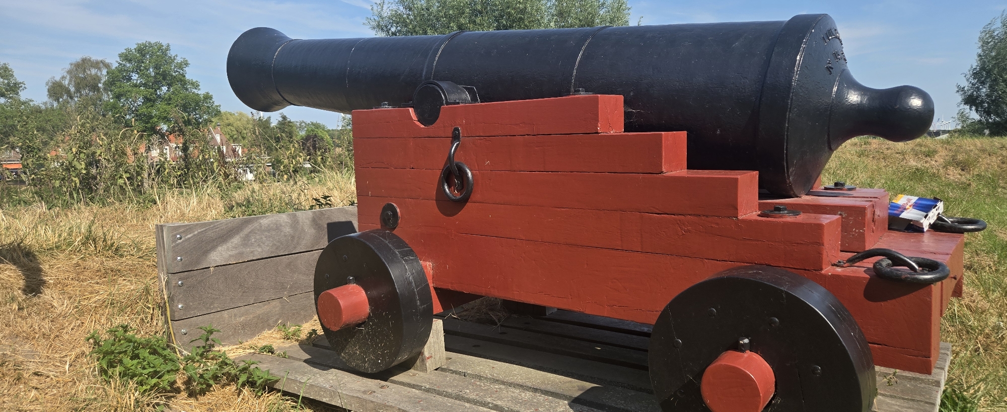 The image shows a close-up view of a historic cannon on a wooden carriage. The cannon is black, and the carriage is painted bright red. The cannon rests on a wooden base, supported by two large black wheels with red hubs. The scene is outdoors, with dry grass and some green vegetation in the foreground and background. In the distance, there are trees and buildings, suggesting a rural or semi-rural setting. The sky is blue with some scattered clouds. A small Red Bull cooler is visible on the right side of the cannon carriage, which is incongruous with the historical setting. There are no people in the image. The location is identified as the city of Brill, Netherlands.