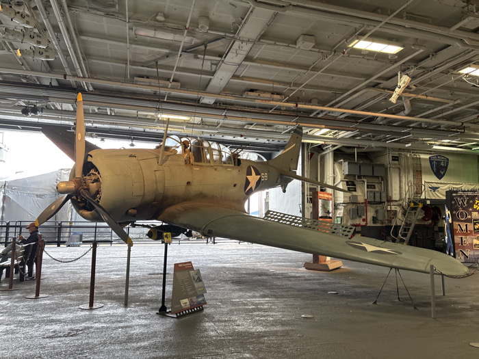 The image shows a World War II-era SBD Dauntless dive bomber on display. The aircraft is painted in a faded green and grey camouflage scheme with white stars. It is supported by stands on a gray hangar deck. A black and white information placard is near the plane. Overhead is a complex network of pipes and supports. In the background, there are stairs and doorways.