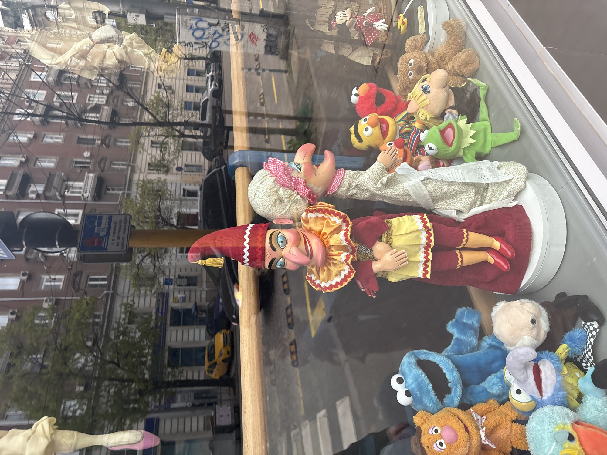 The image displays a vibrant shop window filled with puppets and plush toys, viewed from the exterior, showing significant reflections of the street scene opposite. The picture is rotated 90 degrees clockwise, but the description assumes an upright orientation.
Inside the window display, a variety of cheerful puppets and stuffed animals are arranged. Prominently featured are two large traditional puppets: a male figure resembling a jester with a large nose, wide smile, red and yellow costume, and a pointed hat with a tassel; and a female puppet in a floral dress, white apron, and a pink and white checkered headscarf. Surrounding these, and clustered towards the bottom of the display, are numerous familiar plush characters including Elmo, Cookie Monster, Kermit the Frog, a brown teddy bear, and an orange, bear-like puppet (possibly Fozzie Bear).
The reflections on the glass reveal a typical urban environment. Multi-story buildings with balconies and windows line a street, where parked and moving cars, including a yellow taxi, are visible. Trees with budding leaves stand along the roadside. A pedestrian crossing is faintly discernible on the asphalt. A blue traffic sign with white Cyrillic text is clearly reflected, reading "П 25 ТЕГЛЕНЕ С ОТПАДНАЛИ РАЗРЕШИТЕЛНИ," which translates to "P 25 TOWING WITH EXPIRED PERMITS." Graffiti, including the number "88," is visible on one of the reflected buildings.
The scene takes place during the daytime, likely under an overcast sky, given the soft, diffused lighting and absence of strong shadows. The Cyrillic text on the traffic sign strongly indicates a location in a country where this alphabet is used, aligning with the provided context of Sofia, Bulgaria. The overall impression is a charming, playful shop window integrated into a bustling city streetscape.