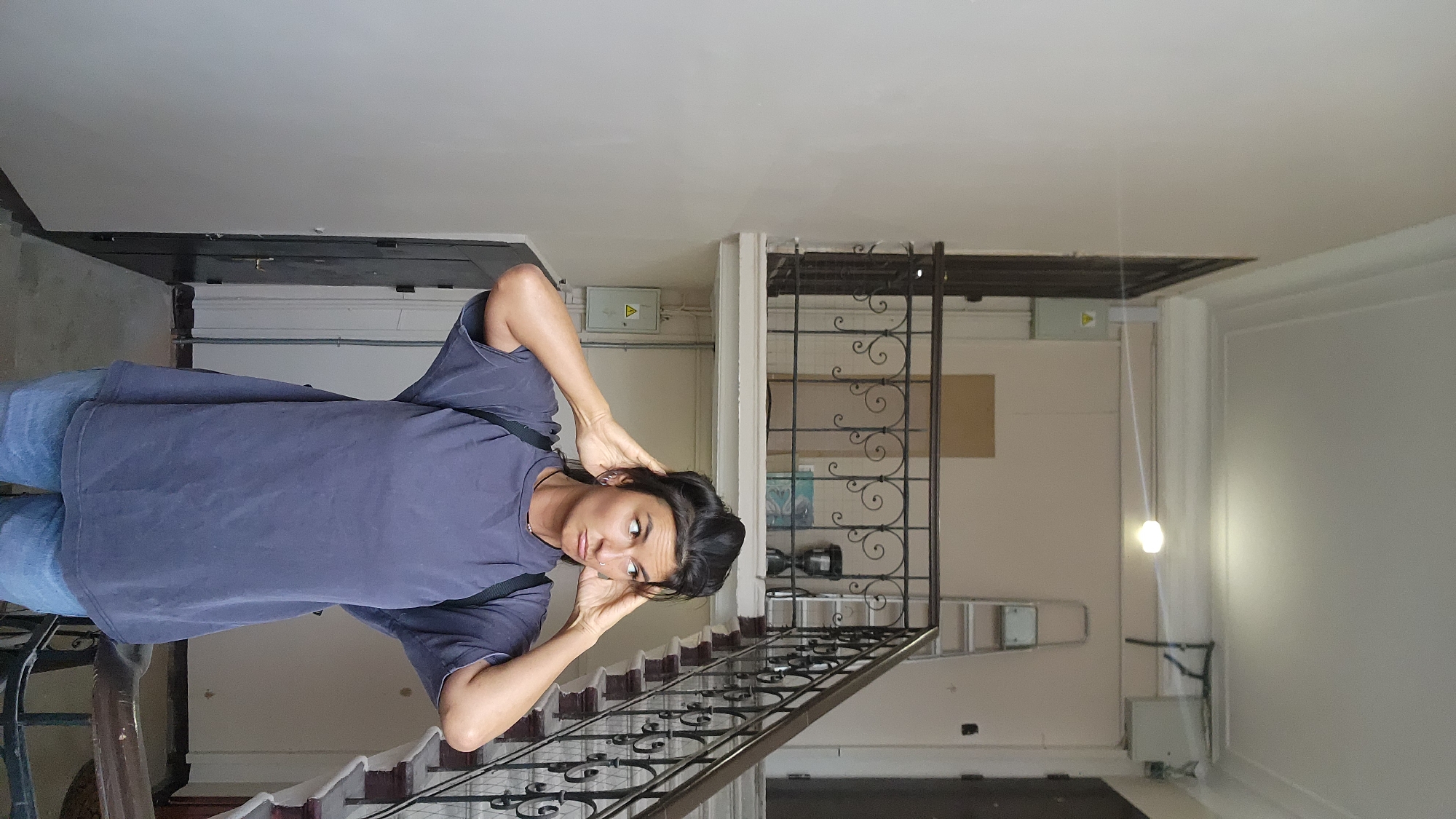 A medium-shot, vertical portrait depicts a person of indeterminate gender on a staircase with ornate iron railings, located inside a building. The person faces the viewer with a questioning look, their hands raised to the sides of their head. They are wearing a dark purple t-shirt and denim jeans. A dark shoulder bag strap is visible across their body.
The staircase leads upwards towards a white ceiling. A metal gate with elaborate scrollwork is visible behind the staircase. A ladder is leaning against the wall behind the gate. A hanging light bulb illuminates the scene on the right. Electrical boxes are mounted on the wall. The location is indicated to be in the city of Moscow, Russia.