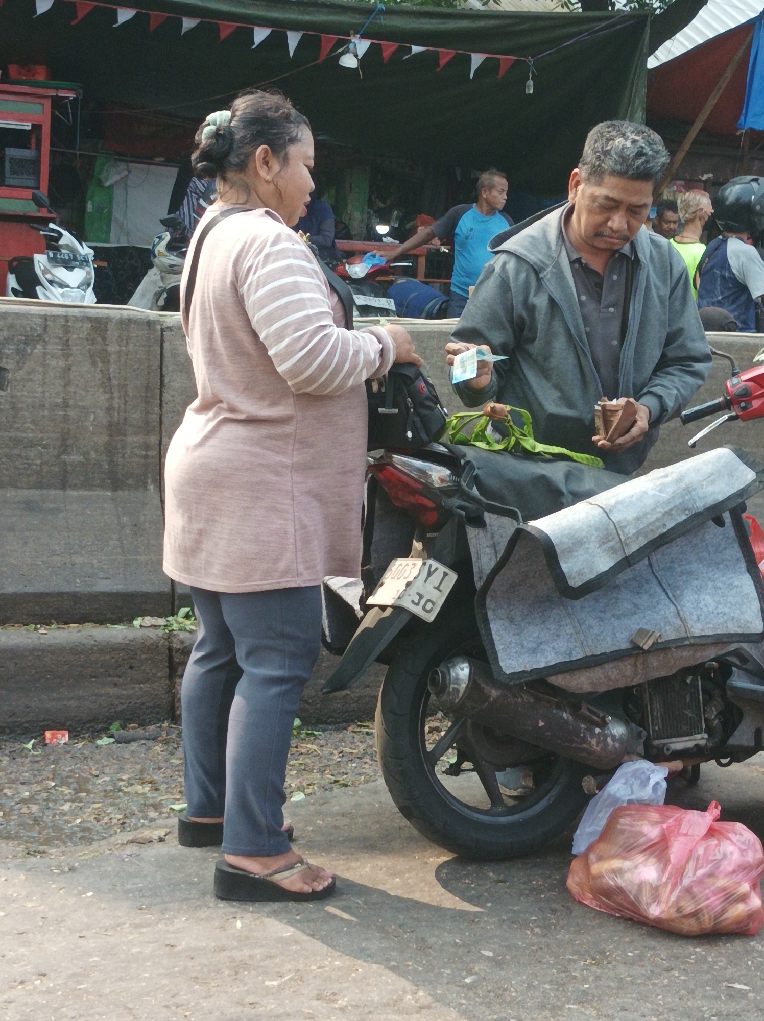 The image depicts a street scene in South Jakarta, Indonesia, with two adults interacting near a motorcycle. A woman wearing a pink and white striped long-sleeved shirt and dark pants stands facing right, her body turned slightly away from the viewer. She is wearing dark wedge sandals. Next to her, a man in a grey hooded jacket and dark shirt is holding out a small piece of paper or currency and a small wooden object. He is looking down at these items. A motorcycle with a light-colored cover on its rear is parked behind them. A red plastic bag filled with what appear to be food items is on the ground next to the motorcycle's rear wheel. In the background, there are stalls or vendors with awnings and some other people are visible. The environment suggests an outdoor market or roadside area with concrete barriers and paved ground.