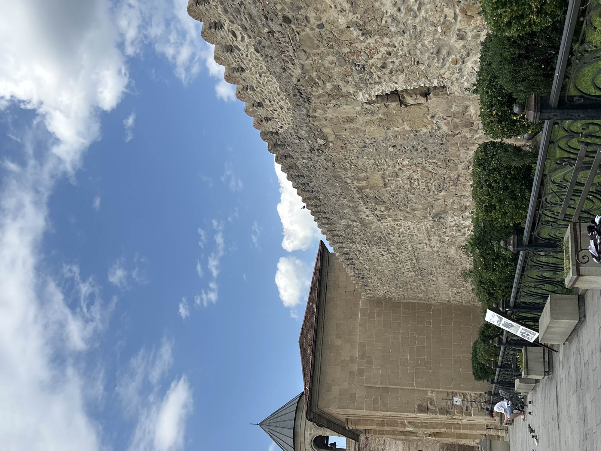 This is a vertical, eye-level, outdoor shot featuring a stone wall and part of a building in Mtskheta, Georgia. The sky above is blue with scattered white clouds. The stone wall, which dominates the right side of the frame, is textured and appears old, with some sections broken or crumbling. The top of the wall has a crenellated design. To the left of the wall is a light-brown stone building with a red-tiled roof. A conical tower with a dark roof and a bell is attached to this building. In the foreground, a paved walkway runs alongside a black metal fence. Behind the fence are trimmed green bushes. A young person in a white shirt and blue shorts is sitting on a bench to the left, with several pigeons gathered around them. A white sign on an easel stands near the bench. The overall scene suggests a historical or religious site.