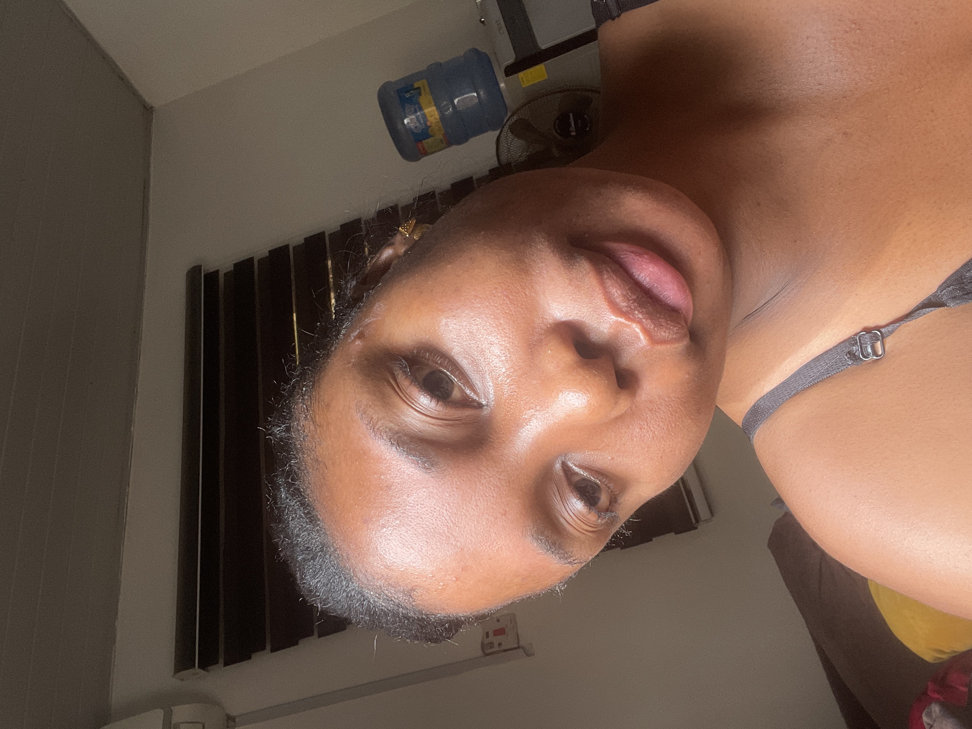 A close-up, dimly lit selfie taken indoors. A dark-skinned woman is the main subject. She faces the camera with a slight tilt of her head. Details of her facial features, including eyes, nose, and lips, are visible. A grey bra strap is visible on her shoulder. In the background are window blinds, a water cooler, and an electric fan. The image was taken at an angle that is not straight. There is no information to conclude if this was taken in Abuja, Nigeria.