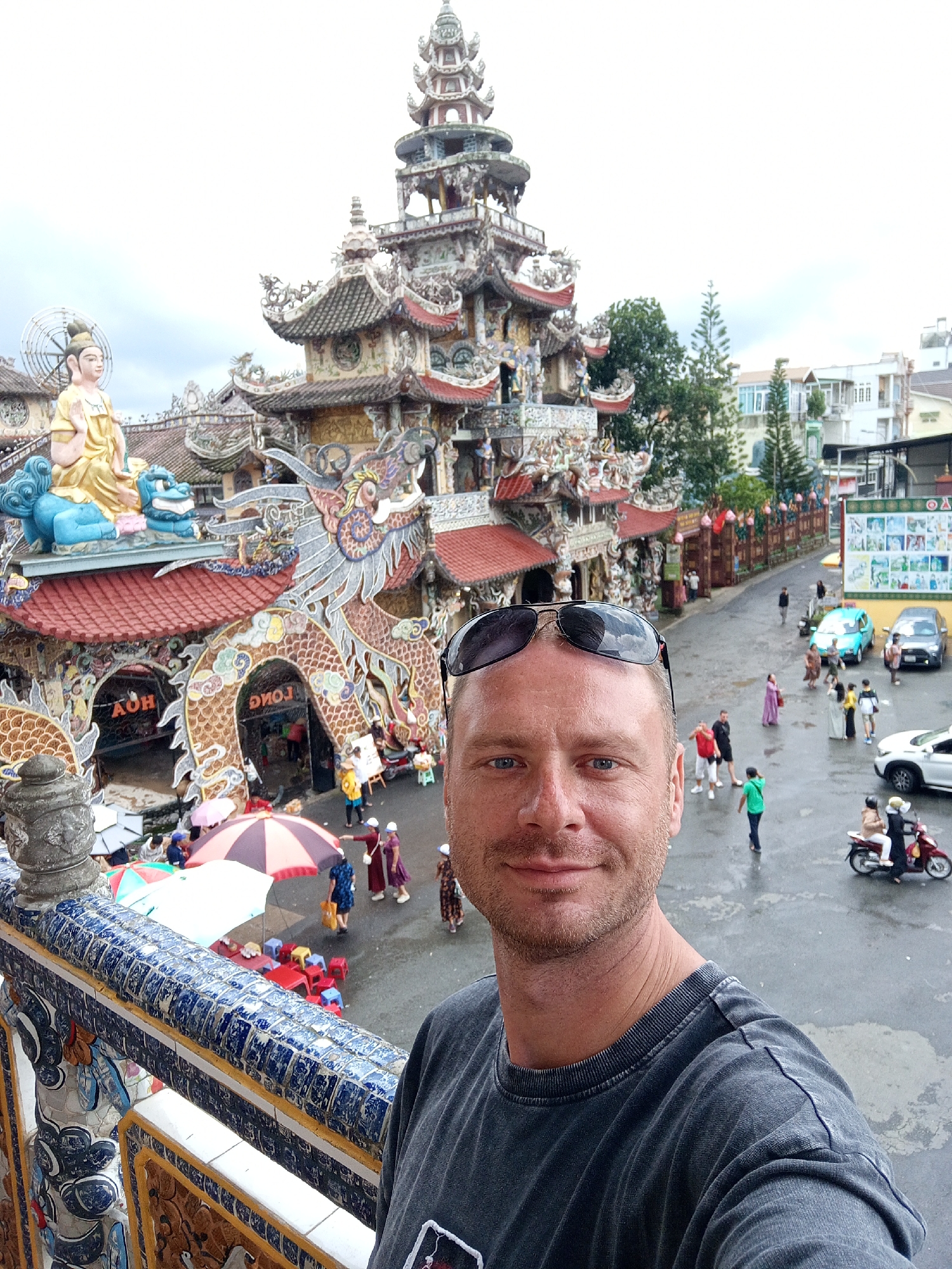 This is a selfie taken by a man with short blonde hair and blue eyes, wearing a dark grey t-shirt and sunglasses. He is standing on a balcony with blue and white tiled railings overlooking a street in Xuân Thành, Vietnam. In the background, there is a large, intricately decorated building with a pagoda-style tower and a dragon sculpture. Several people are walking on the wet street, some carrying umbrellas. There are also several vehicles, including cars and motorcycles. The sky is overcast.