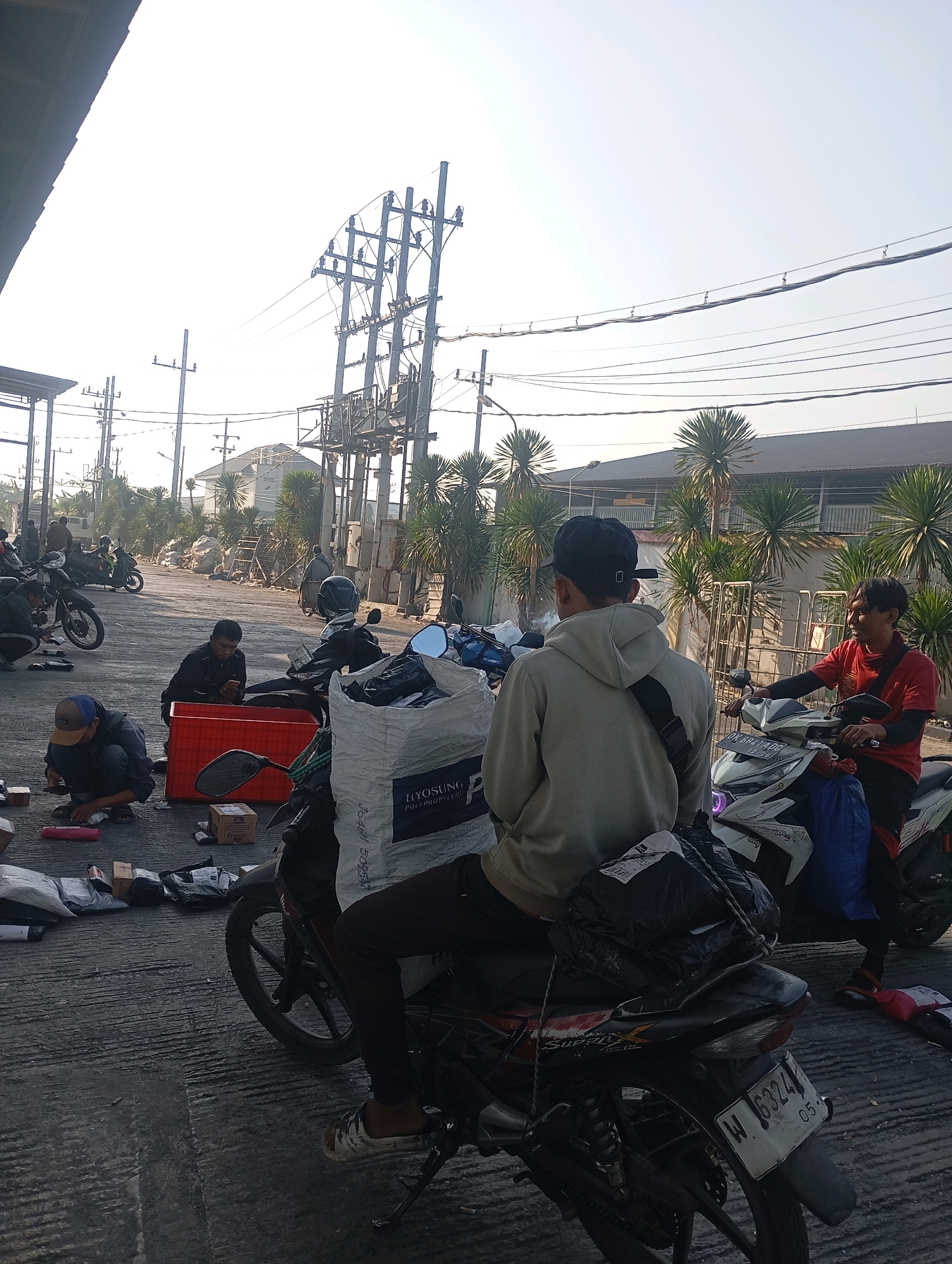 A group of people are gathered around motorcycles and packages in what appears to be an outdoor logistics or delivery area in Jabaran, Indonesia. The scene is likely a sorting or dispatch point for goods. The ground is paved and has visible tire tracks and markings. In the background, there are palm trees, buildings, and utility poles with numerous wires, typical of an urban or semi-urban Indonesian environment. The sky is bright and appears to be daytime, with hazy sunlight.
Several individuals are actively engaged in tasks related to the packages and motorcycles. One person, wearing a red t-shirt and a black helmet, is seated on a white motorcycle, looking towards the viewer and smiling. Another individual, wearing a grey hoodie and a black baseball cap, is also seated on a motorcycle, facing away from the camera. They appear to be interacting or communicating.
Other people are seen crouching or kneeling on the ground, handling packages of various sizes, some of which are wrapped in plastic or paper. One person is looking at a mobile phone while sitting next to a red plastic bin. A large white bag with text is prominently placed on one of the motorcycles, possibly containing more items. Several motorcycles are parked closely together, indicating a busy operation. License plates on the motorcycles are visible, with one reading "W 6324". The overall atmosphere suggests a functional, perhaps informal, distribution hub.