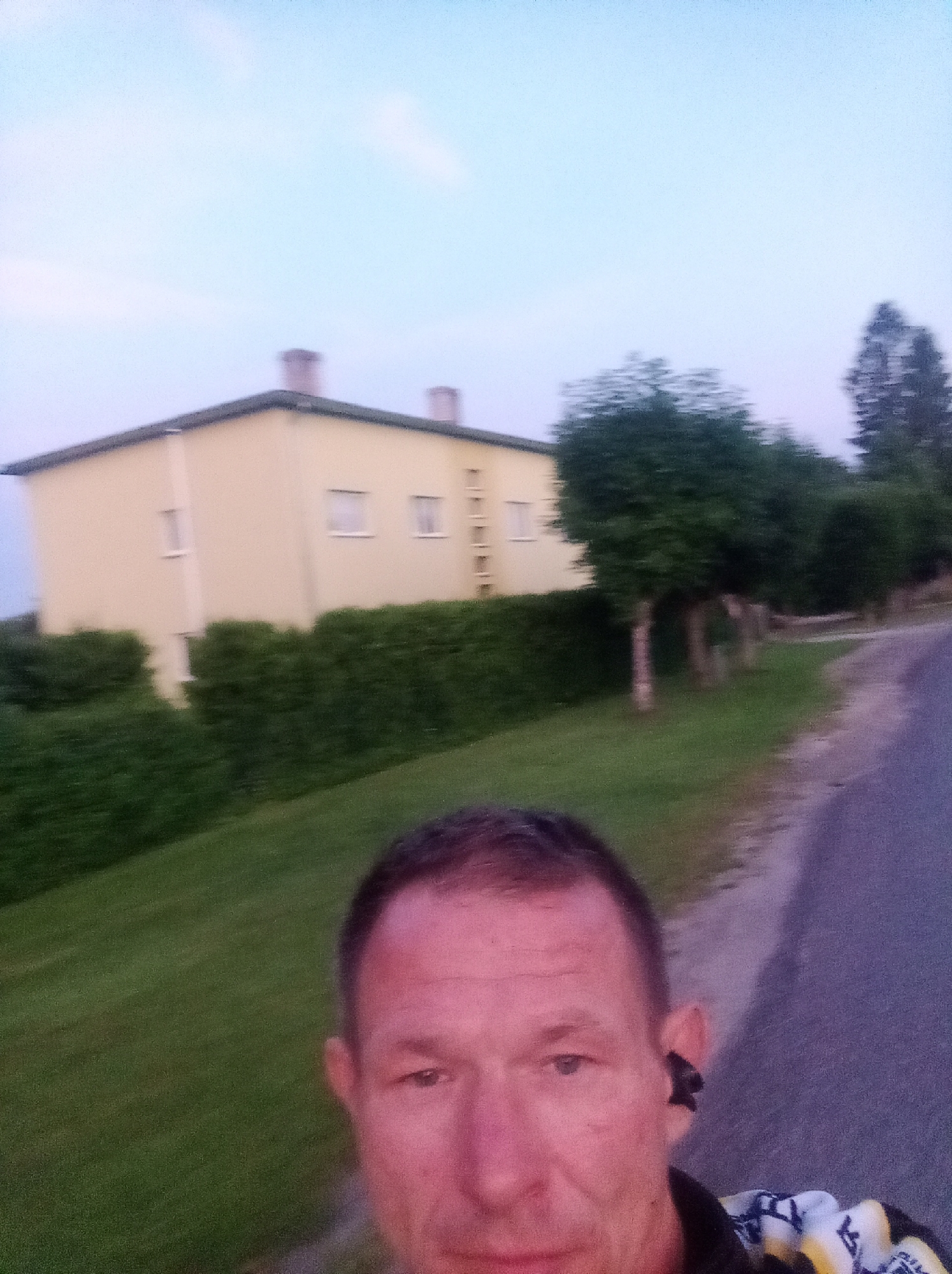 In Tapa, Estonia, a man is captured in a selfie while standing next to a paved road. He is wearing a yellow and white patterned garment and black wireless earbuds. In the background, a yellow building with a dark roof and chimneys stands behind a row of green bushes. Several trees are visible, some with visible trunks, and a large coniferous tree is to the right of the building. The sky is a soft blue. The scene suggests an outdoor setting, possibly a residential area, during either dawn or dusk, given the ambient light conditions. The image is somewhat blurry, indicating movement or a lower resolution.
