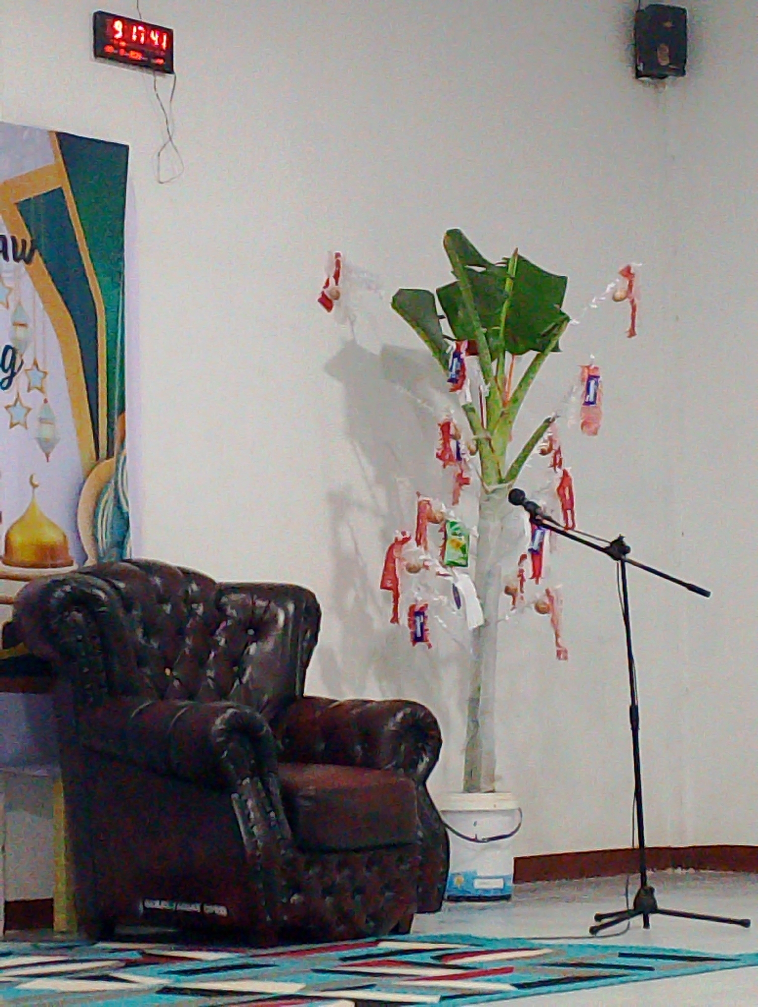 The image depicts an indoor scene, possibly a hall or room, with a dark brown leather armchair prominently placed on the left. To the right of the armchair, a young banana tree is visible in a white bucket, adorned with small, colorful, wrapped items. A black microphone stand with a microphone is positioned in front of the banana tree. The background consists of a plain white wall. On the wall, a digital clock displays "9 17 41", and a dark speaker is mounted high up on the right side. A banner with Islamic motifs, featuring a golden dome and lanterns, is partially visible on the far left. The floor is covered with a colorful rug. There are no people in the image. The location is specified as Bobong, Indonesia.