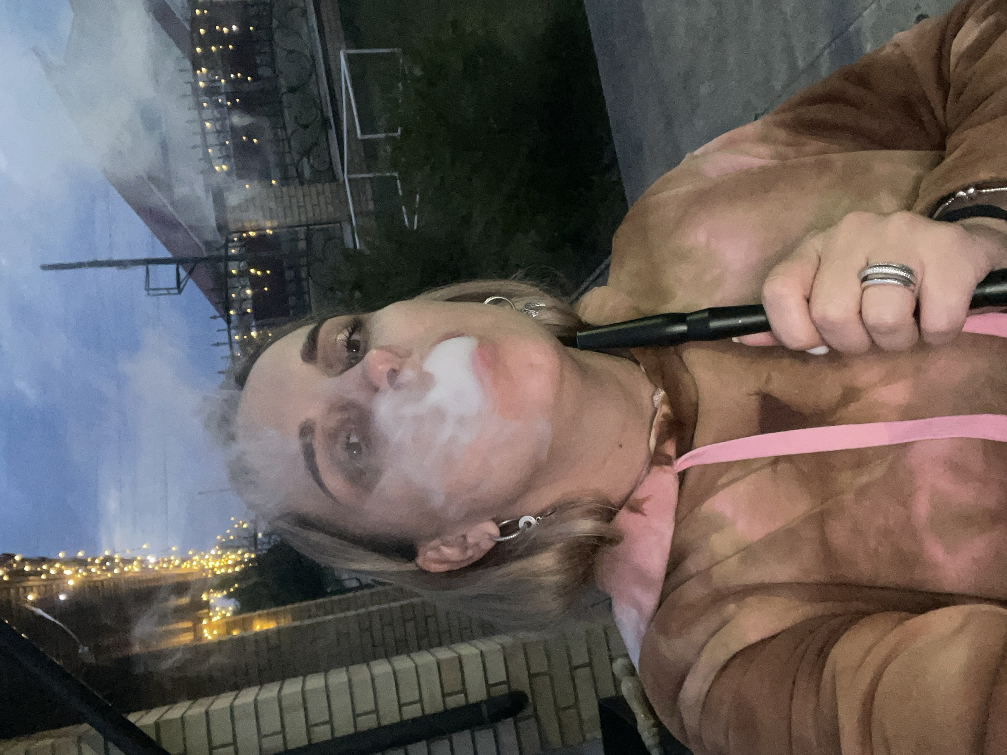 A woman with blonde hair and fair skin exhales a cloud of smoke, likely from a hookah pipe she holds to her lips. She wears silver hoop earrings and multiple rings on her left hand. Her clothing is a brown and pink tie-dye pattern. In the background, a fence made of light-colored brick can be seen. Beyond the fence, strings of yellow lights illuminate a structure, possibly a building or a street, giving a cityscape view. The sky is a darkening blue, suggesting it is evening. The location is Chaikovsky, Russia.